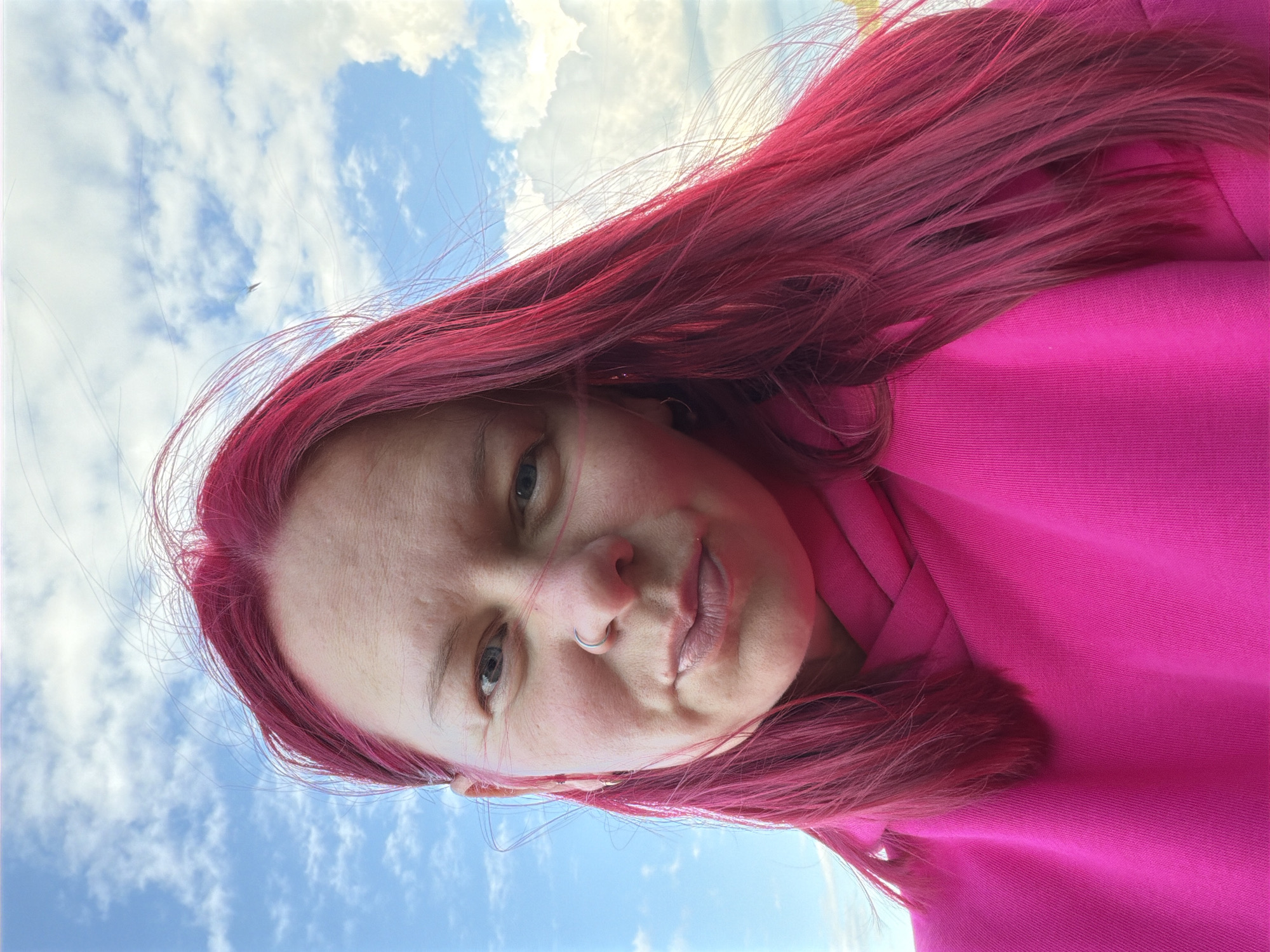 A close-up, low-angle portrait depicts a person with vivid bright pink or magenta hair, looking directly at the camera. They are wearing a matching pink hooded garment. A silver hoop nose ring is visible on their left nostril. The background features a bright blue sky with scattered white, fluffy clouds. A small, indistinct object, possibly an airplane or bird, is discernible in the upper left area of the sky. The lighting suggests a clear, sunny daytime. The person's expression is neutral and composed. The original image is oriented sideways, giving the impression of an upward-looking shot. This scene is located in Chaikovsky, Russia. No visible text is present.