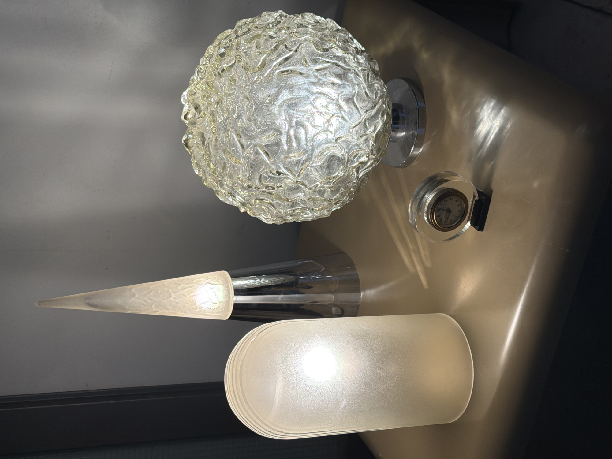 The image shows an arrangement of objects on a surface. A textured glass globe sits atop a chrome base. To its right is a small analog clock encased in clear plastic. Below the globe is a chrome-and-frosted-glass cone-shaped lamp, positioned above a larger cylindrical frosted glass lamp. The surface appears to be tan. The location in Buenos Aires, Argentina, and details about people or interactions are unknown.