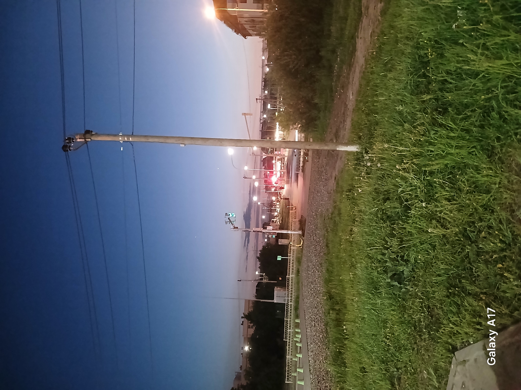 The image depicts an urban landscape at dusk in Yekaterinburg, Russia. Power lines extend from a utility pole in the foreground against a blue and pink sky. To the right is a stretch of green grass and a dirt path. In the distance, a street with traffic lights shows red brake lights, indicating vehicular movement. Buildings and streetlights dot the background. There are traffic lights showing green color.