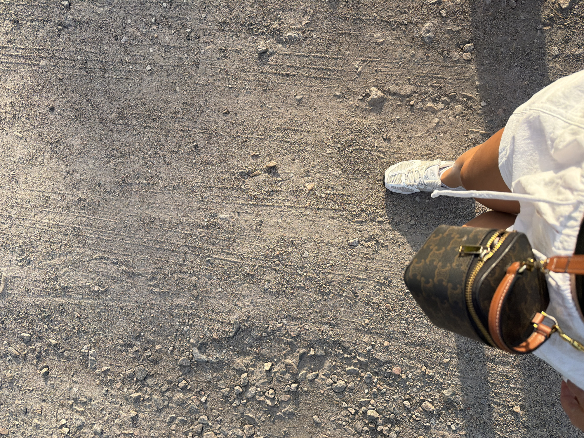 This media file captures a ground-level perspective of a person's leg and arm, holding a small cylindrical handbag. The person is wearing a white garment, possibly shorts or a skirt, and a white sneaker with a tan sole. The handbag is dark brown with a repeating pattern of dark brown and tan, featuring gold-toned hardware including a zipper and clasps. The bag is worn on a brown leather strap.
The scene is set on a surface of gravel and dirt, with visible tire tracks and scattered small stones. The lighting suggests it is daytime, with long shadows cast on the ground, indicating either early morning or late afternoon. The overall impression is casual and outdoors. The location is identified as Dnipro, Ukraine. There is no visible text in the image.