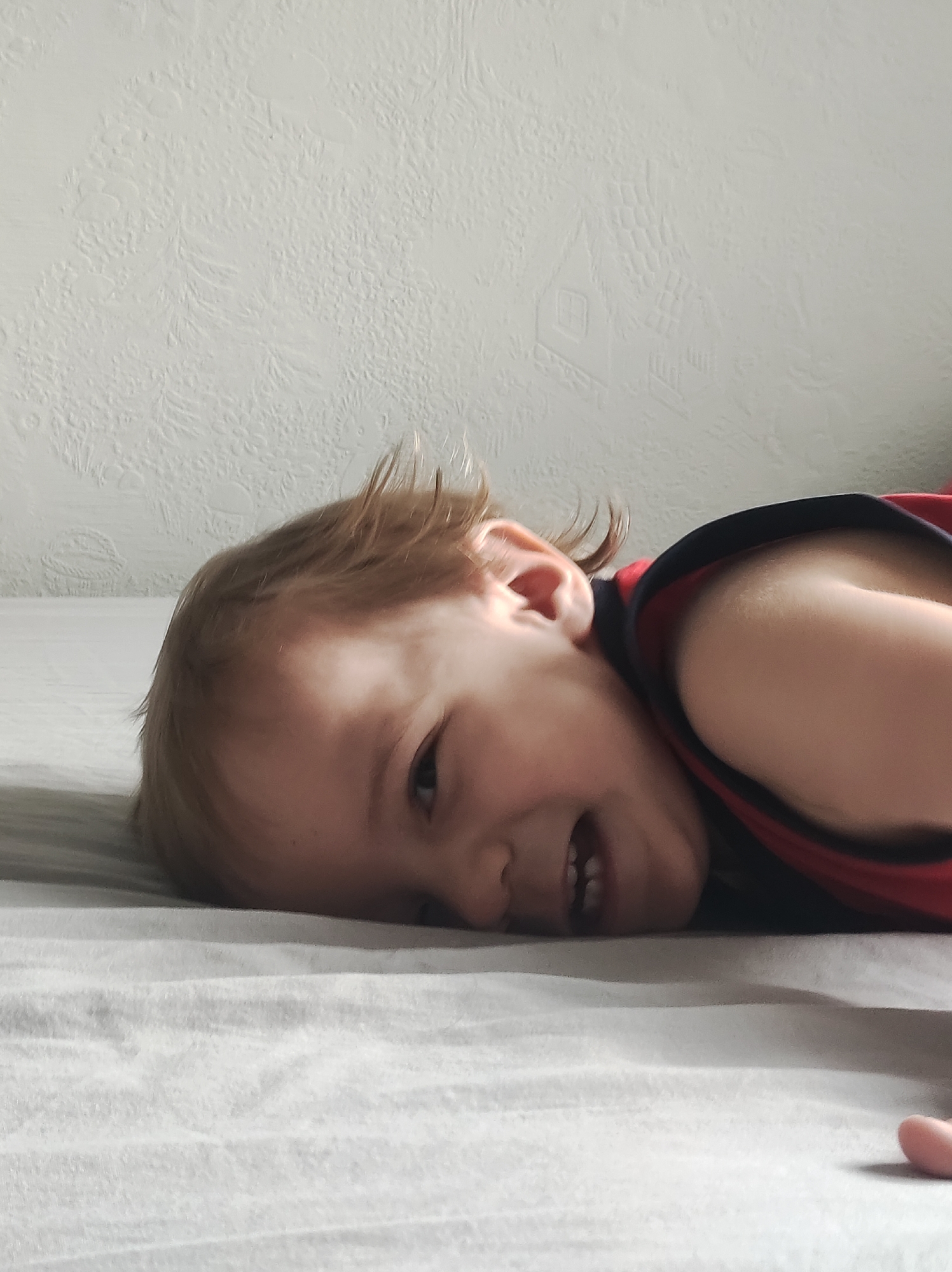 A young child is lying on a bed with a gray sheet. The child is wearing a red and black tank top. The background shows a white wall with a faint patterned design. The child is smiling. The location is Chaikovsky, Russia.