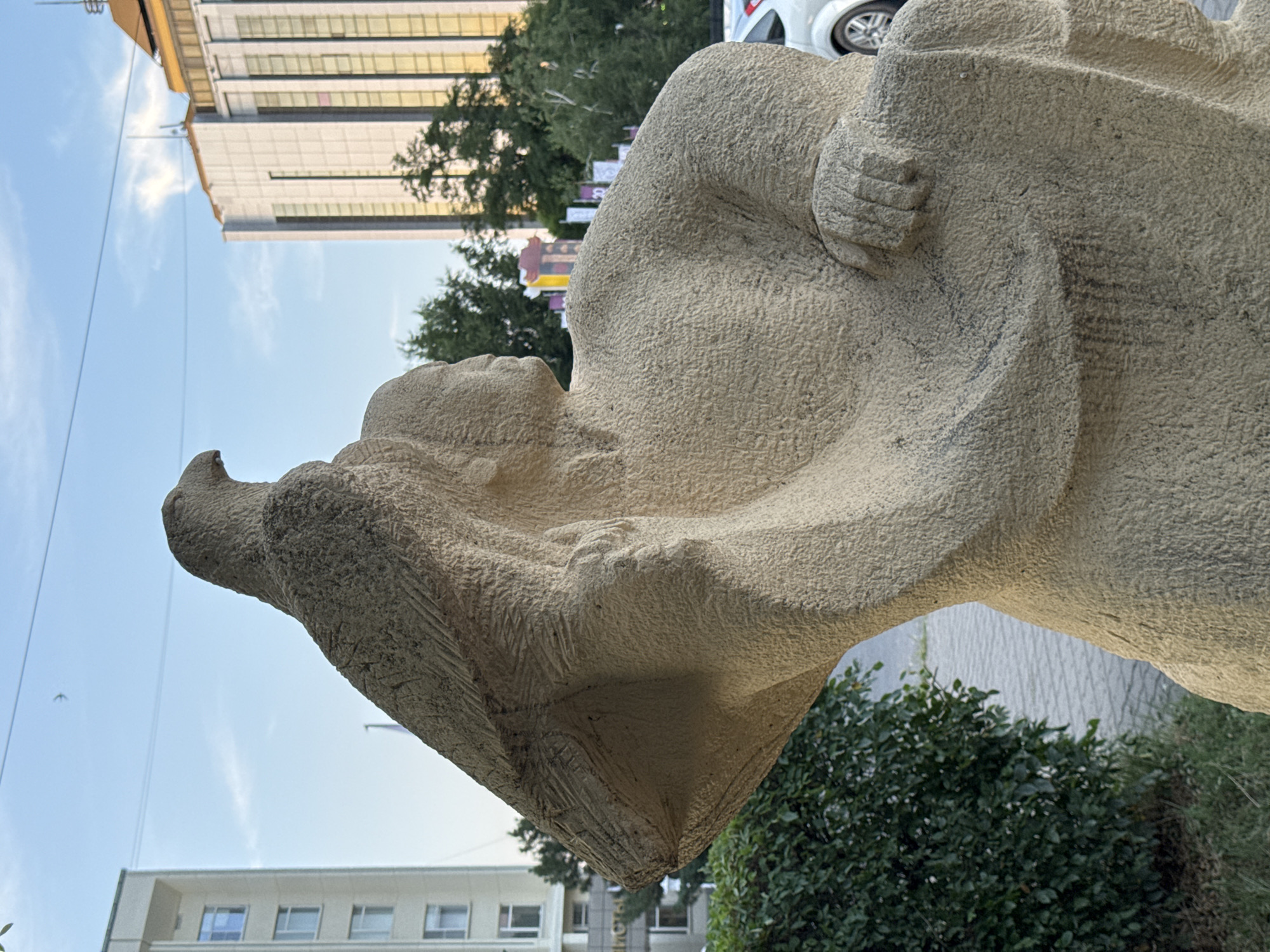 The image shows a stone sculpture of a man with an eagle perched on his shoulder. The sculpture is made of a rough, textured material and has a weathered appearance. The man's body is depicted in a dynamic pose, with his head turned to the right, looking towards the sky. The eagle is carved with detailed feathers and appears to be in mid-flight.
The sculpture is situated in an urban environment. In the background, there are modern buildings with glass facades and a golden-colored roof. Trees and some greenery are also visible, suggesting a park or public space. The sky is a clear blue with a few wispy clouds. The lighting suggests it is daytime, possibly late afternoon or early morning, as the sunlight casts soft shadows.
This sculpture is located in Elista, Russia, the capital city of the Republic of Kalmykia. Elista is known for its unique Buddhist culture and its numerous chess-themed attractions, as well as its distinct architectural style that blends Soviet-era buildings with more contemporary designs. The presence of a sculpture featuring an eagle, a symbol of power and freedom, could be interpreted in various cultural contexts within Russia and Central Asia. The surrounding buildings suggest a central or administrative area of the city.
