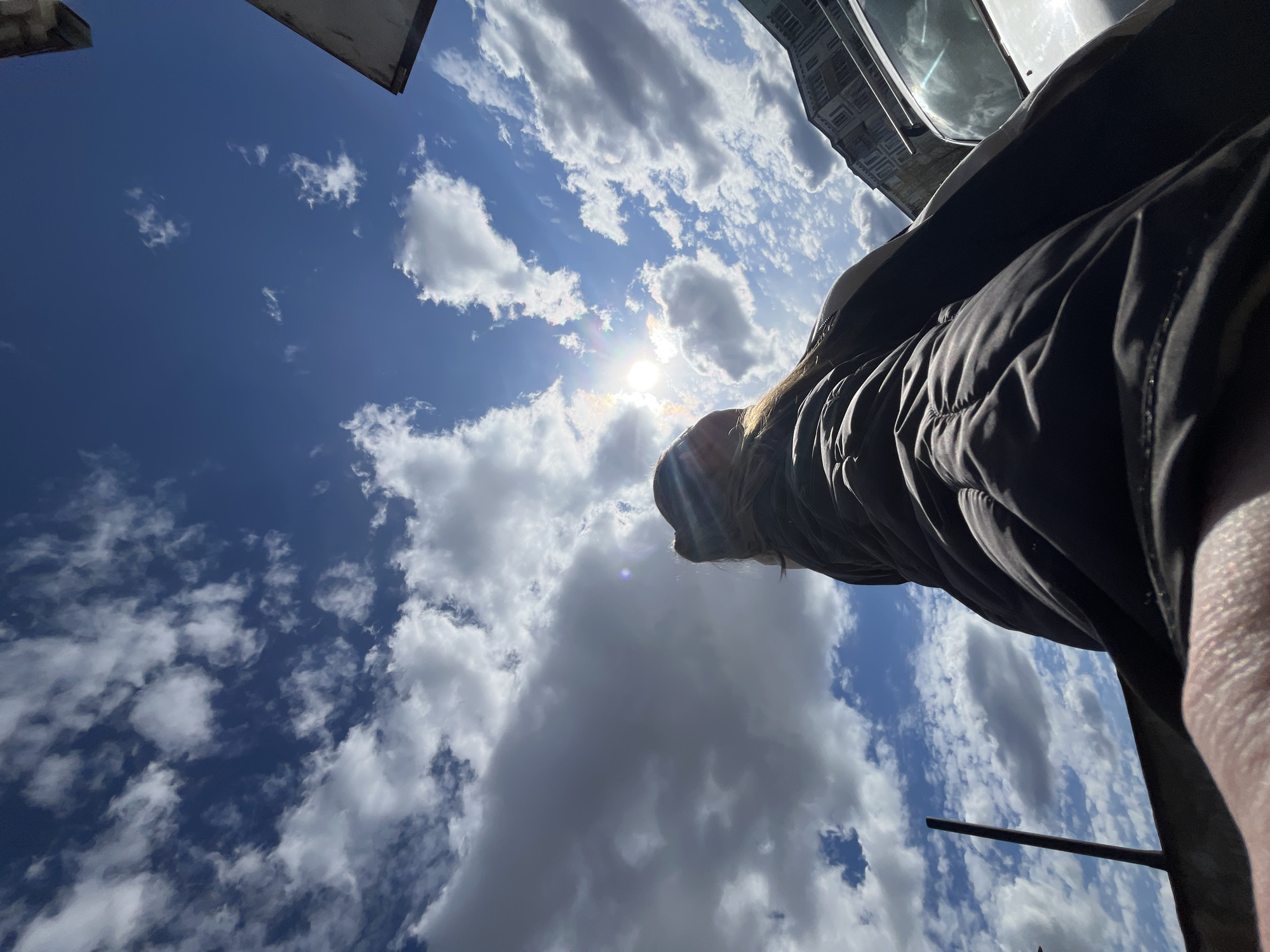 The image is taken from a low angle, looking upwards towards a partially cloudy sky. The sun is visible, obscured by some clouds, causing lens flare. A person wearing a dark jacket is in the foreground, partially blocking the view of the sky. Building tops are visible in the top left and right corners of the image. The location is possibly Vysokovsk, Russia based on the prompt.