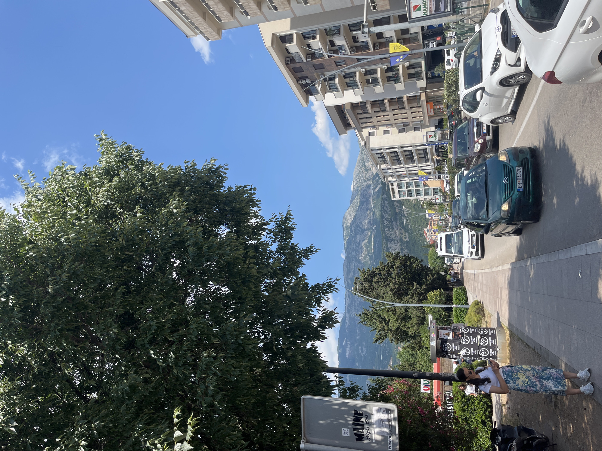 This is a vertical, eye-level, outdoor shot in what appears to be a bustling city environment, likely in Budva, Montenegro, given the landscape and architecture. The day is bright and sunny, with a clear blue sky dotted with a few wispy clouds. The scene is dominated by a dense, leafy green tree on the left, partially obscuring the view.
In the midground and background, modern buildings with multiple stories and balconies line a street where a line of cars are stopped, suggesting traffic or a busy urban area. Among the vehicles, a green Citroën with license plate "BR DA 370" is visible, as is a white Toyota Yaris. A woman in a white top and a patterned skirt stands on the sidewalk, looking at her phone, with a motorcycle parked behind her. Adjacent to her is a kiosk or stand with posters and signage, including text that reads "MAINE Night Club". The backdrop features a scenic mountainous landscape with terraced slopes, hinting at a coastal or mountainous region. The overall atmosphere is one of everyday urban life on a pleasant day.
