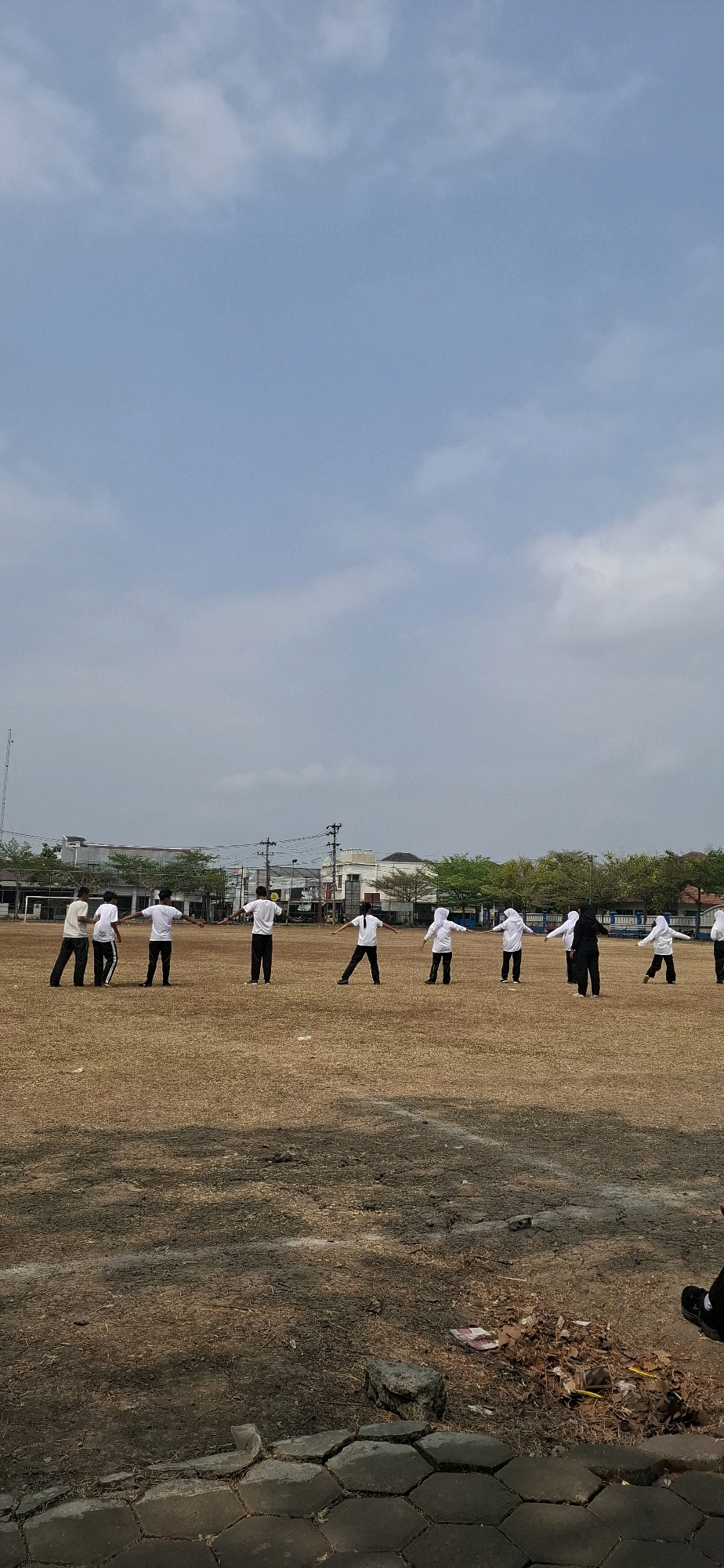 In Baturan, Indonesia, a group of approximately ten individuals, dressed in white shirts and black pants, are gathered on a dry, grassy field. Most are wearing white hijabs. They are arranged in a semi-circle and appear to be performing synchronized exercises, with their arms outstretched to the sides. In the background, several buildings with tiled roofs are visible, along with trees and utility poles. The sky is overcast with scattered clouds. The ground is uneven and covered in dry grass, with some scattered debris and a paved border in the foreground.