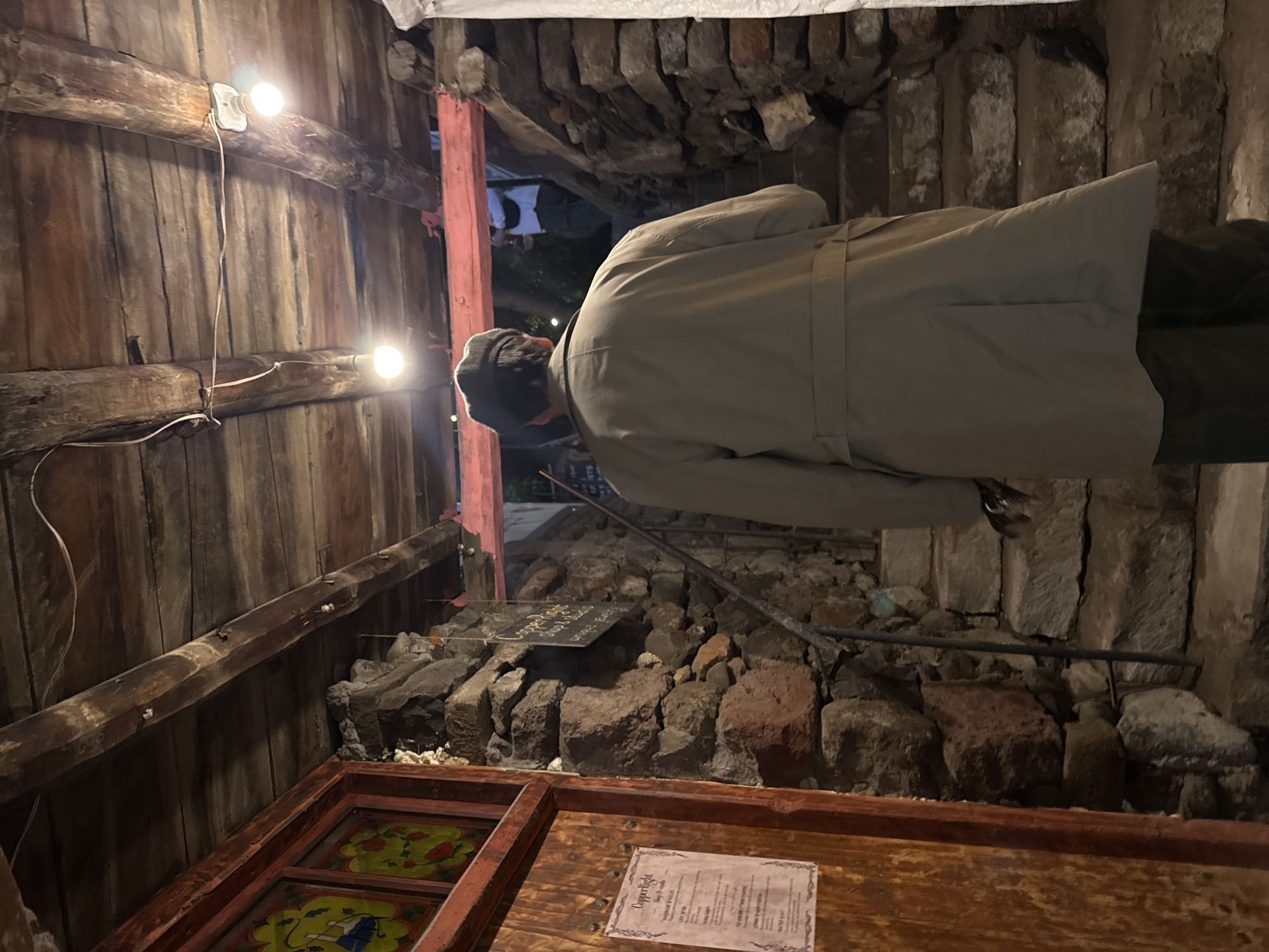 A person wearing a light gray trench coat and a dark cap stands indoors, presumably in Yerevan, Armenia. The setting features a rustic, possibly historical or stylized environment. The foreground consists of wooden structures with painted decorations and a menu card. Mid-ground has a stone wall or foundation with a sign. Background reveals an outside view, possibly an alley or street. The lighting includes exposed light bulbs on the wooden wall, contributing to a warm, aged ambiance.