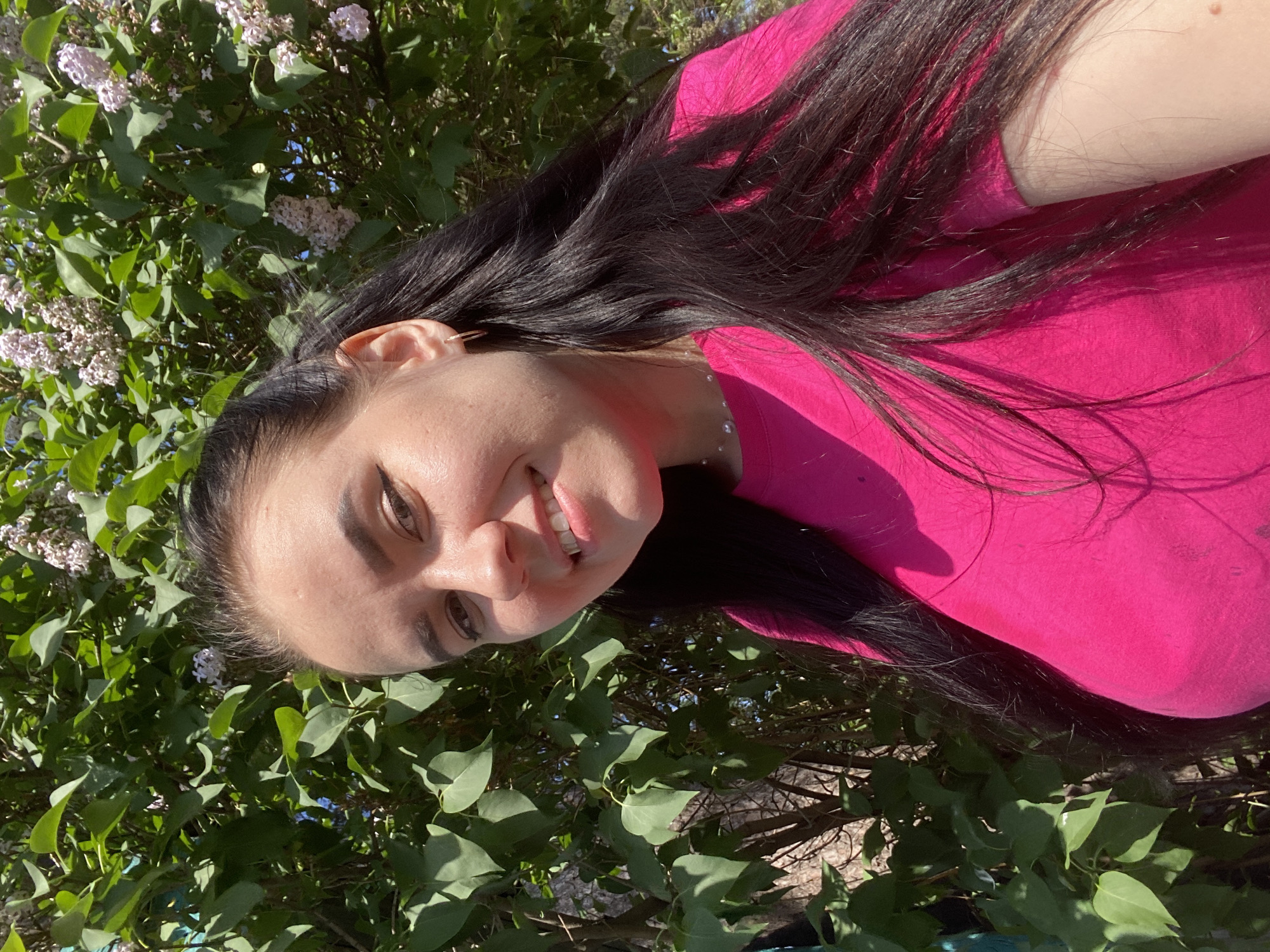 A woman with dark hair and a pink shirt is posing for a picture in front of a lilac bush. The photo was taken outdoors in Chaikovsky, Russia.
