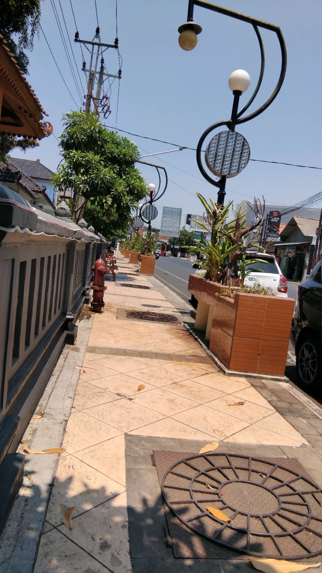 The image depicts a street scene in Batang, Indonesia. The perspective is from a sidewalk looking down the street. On the left, there is a grey fence with vertical slats, and a red fire hydrant is positioned near it. The sidewalk is paved with light-colored tiles arranged in a diamond pattern. There are several manhole covers on the sidewalk. Decorative planters with plants are placed along the edge of the sidewalk bordering the street. Streetlights with spherical white lamps and decorative metalwork are visible. A white car is parked on the street. Buildings and utility poles are present in the background. The sky is clear and blue. The scene appears to be a public area, possibly a commercial district, with infrastructure for utilities and street lighting.