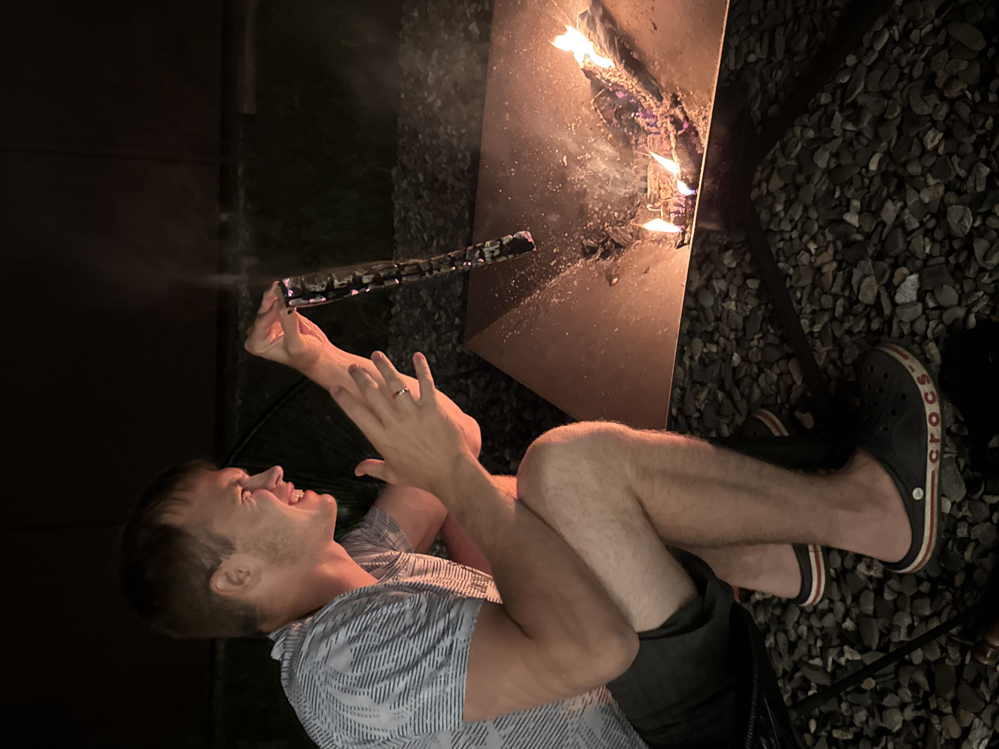 A man, wearing a patterned t-shirt and dark shorts, is sitting on a chair next to a fire pit at night. He is holding a stick over the fire, which is burning wood and embers. The man is smiling and appears to be engaged in an activity with the fire. The surroundings are dark, with gravel on the ground. The image appears to be taken in the city of Starokorsunskaya, Russia.