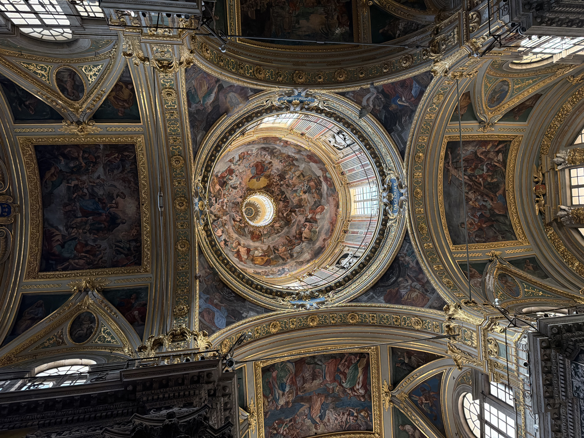 The image shows the interior ceiling of a building, specifically the Basilica della Santissima Annunziata del Vastato in Genoa, Italy. The ceiling is highly decorated with intricate gold trim outlining panels of painted murals, portraying religious scenes with figures. A central dome with a circular opening at the top allows natural light to illuminate the interior. The architecture includes arches, rectangular panels, and triangular segments, all adorned with ornate detailing. Windows are visible in the surrounding structure.