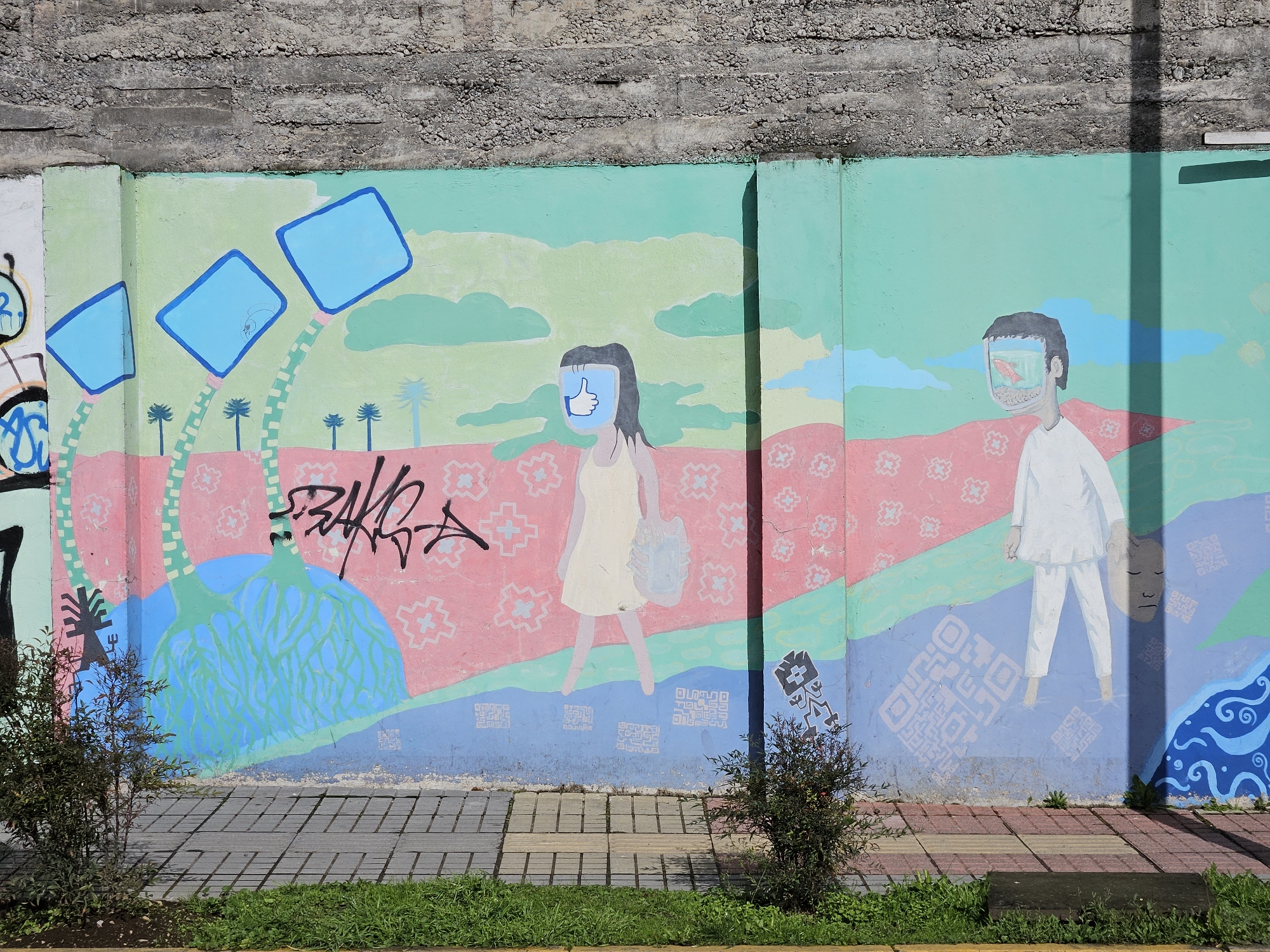 The image is a photograph of a mural on a wall in Osorno, Chile. The mural is divided into two sections. The left section depicts a figure with a blue square head and a "thumbs up" icon on its face. The figure is wearing a light-colored dress and carrying a bag. The background of this section features blue square shapes resembling screens with palm trees. The right section depicts a figure with a similar blue square head filled with an image. The figure is wearing white clothing. The background includes blue cloud shapes. The bottom of the mural has a blue section with white QR codes. Graffiti covers part of the mural. The wall has a rough concrete top. Bushes grow in front of the wall, and a sidewalk with grass separates the mural from the street.