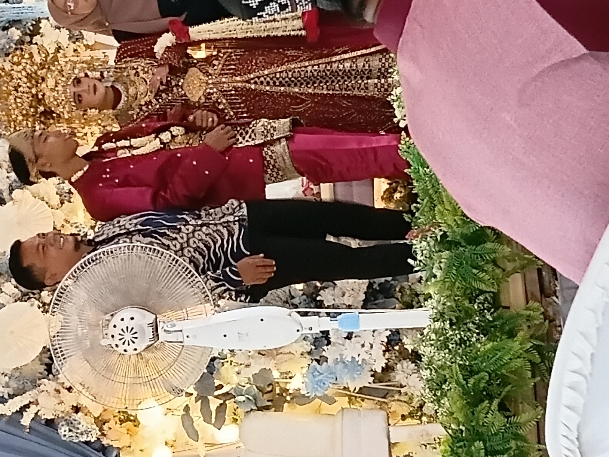 In Sumber Mulyo, Indonesia, at what appears to be a wedding reception, a man in a black and white patterned shirt and black pants stands next to a large white oscillating fan. To his right, a young man in a red suit and traditional headdress stands beside a woman in an elaborate red and gold wedding gown, also wearing a large, ornate gold headdress. The background is decorated with white and blue floral arrangements and shimmering fabric. In the foreground, a person wearing a pink garment is partially visible, seated. The setting suggests a cultural event with traditional attire and celebratory decorations.