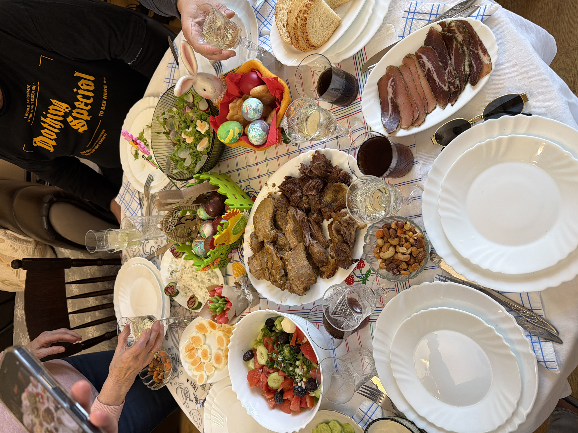 The image shows a dining table laden with food, likely for a festive occasion. The table is covered with a checkered tablecloth. Several dishes are visible: a platter of sliced meat, a bowl of salad with tomatoes, cucumbers, and olives, a plate of boiled eggs, a plate of roasted meat, a bowl of mixed nuts, and bread. A basket of decorated Easter eggs and a small bunny decoration are also present. Glasses of red and white wine, as well as empty crystal glasses, are placed around the table. Sunglasses rest on a plate. Two people are partially visible; one in a black shirt that reads "Nothing Special," holding a glass, and another person holding a smartphone, possibly taking a photo of the table. The specific location in Plovdiv, Bulgaria, cannot be determined from the image alone.