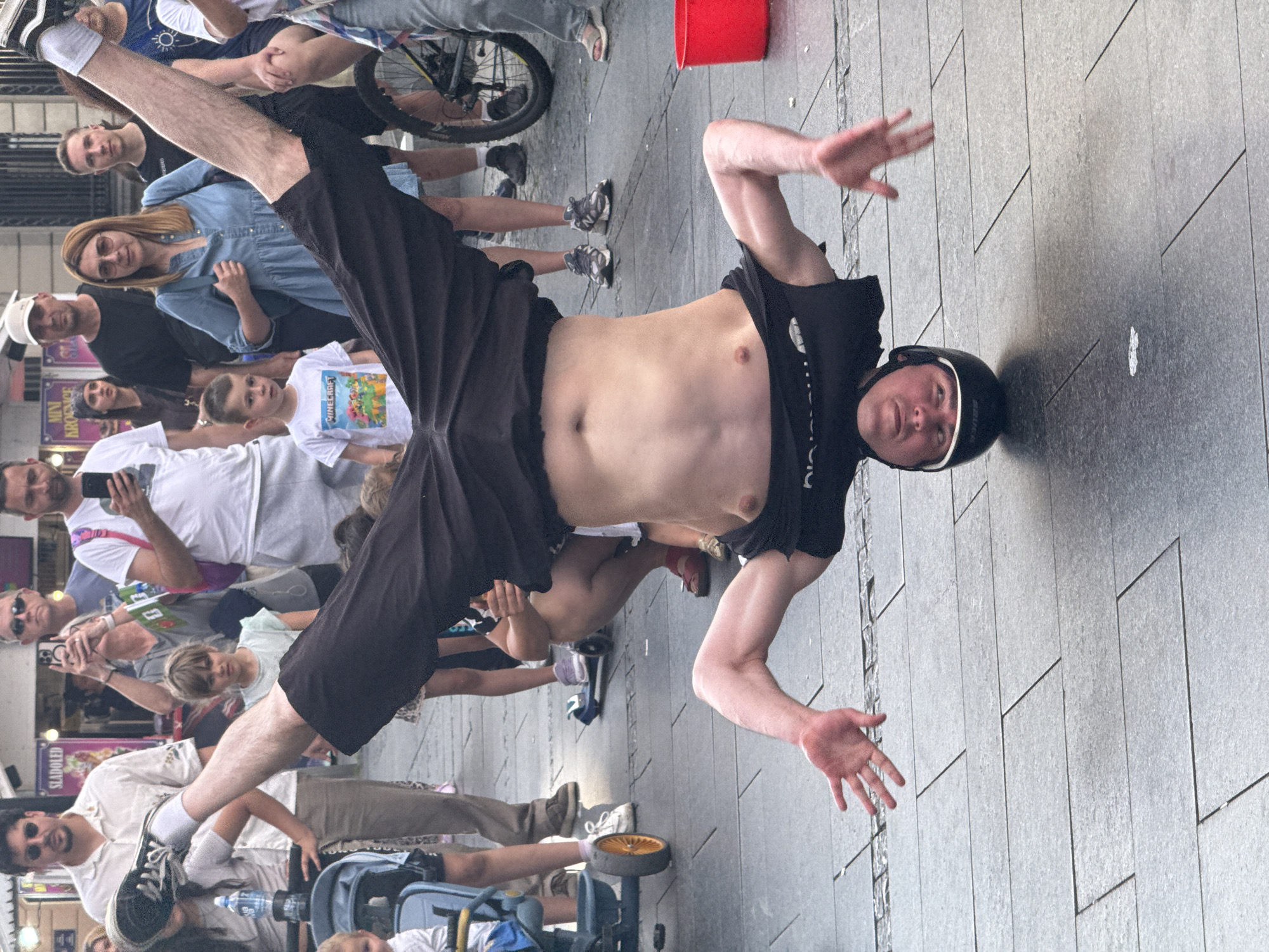 An upside-down man in a black helmet and black shorts is performing a breakdance move on a paved street in Belgrade, Serbia. His hands are on the ground, and his legs are in the air. A crowd of people, including adults and children, watches him. Some people are holding phones to take pictures. A red bucket is visible on the right side of the image. The street is made of large gray pavers. In the background, there are buildings and stalls.
