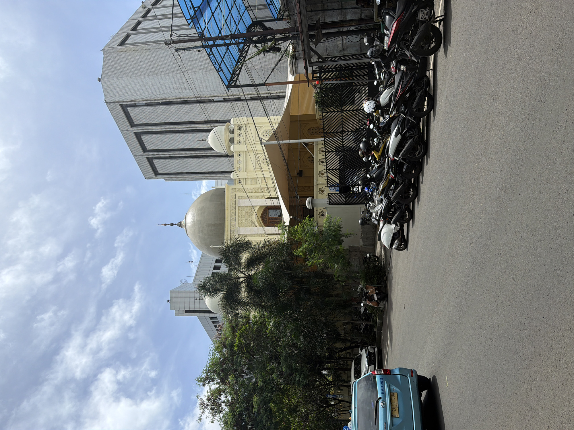 The image shows a street scene in South Jakarta, Indonesia. A mosque with a large dome is prominent, flanked by modern buildings. A row of motorcycles is parked along the street. A blue car is visible at the bottom of the frame, parked along the roadside. Trees and power lines are visible overhead. The sky is partly cloudy.