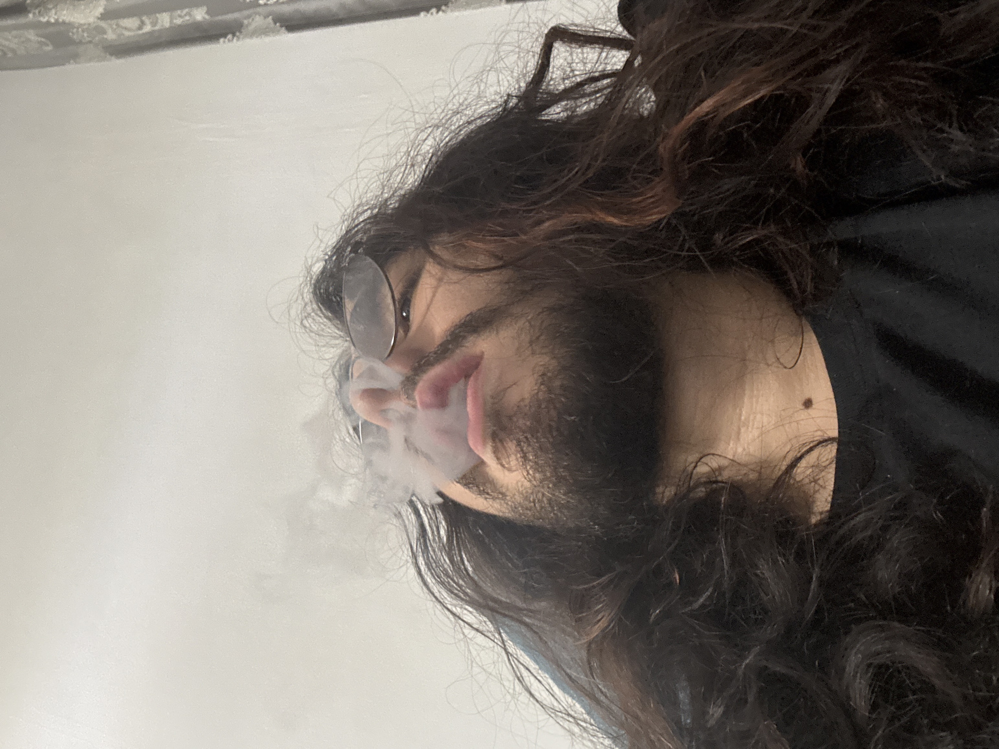 A low-angle, medium shot captures a person with long, dark, wavy hair and a beard exhaling a cloud of smoke. The individual wears glasses and a black t-shirt. The background consists of a plain, lightly textured wall and a section of a patterned curtain on the right side of the frame. The lighting is soft, casting subtle shadows.