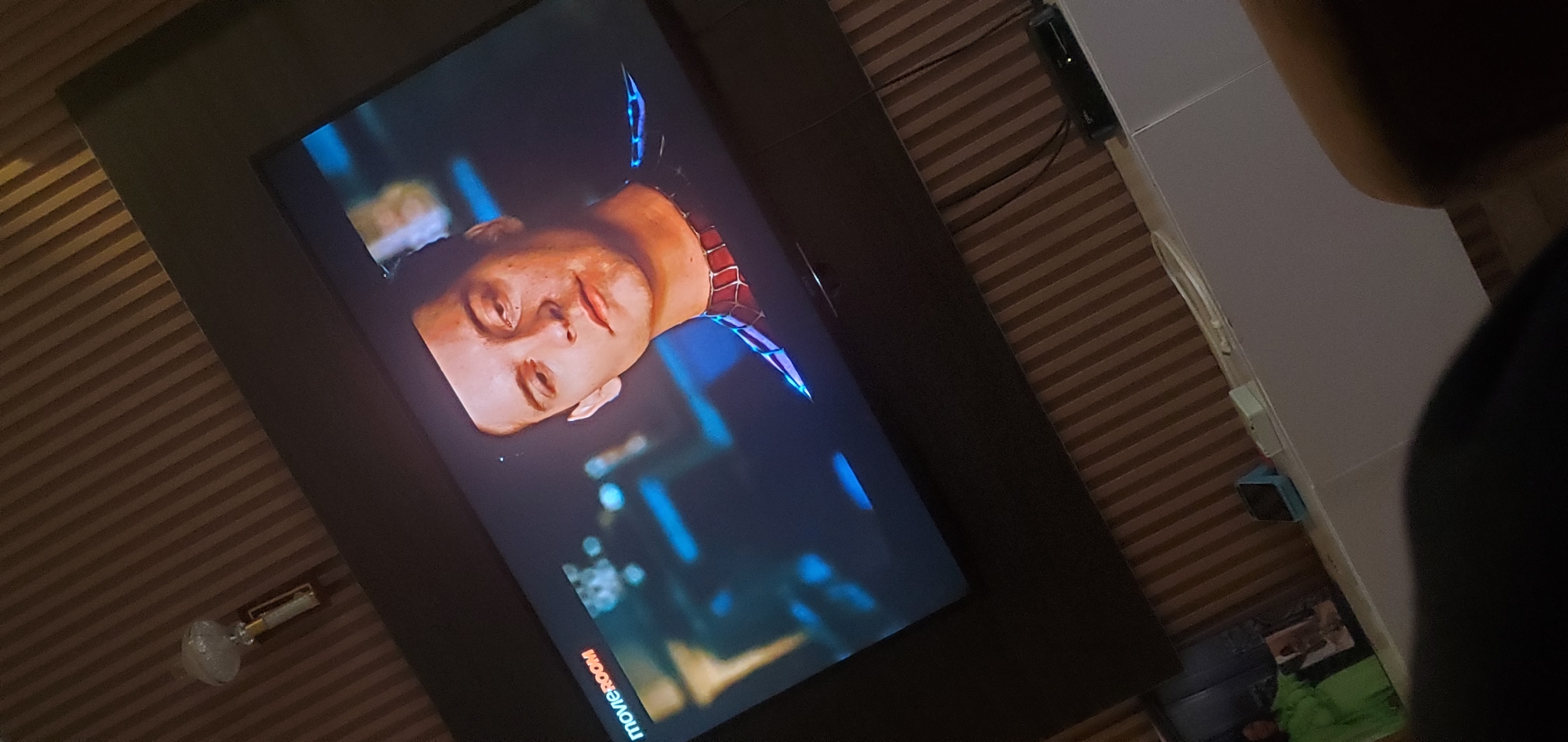 The image shows a close-up view of a television screen displaying a scene from a movie, likely a superhero film. The primary subject on screen is a person's face, tilted upwards. The individual appears to be wearing a red and blue suit with a web-like pattern, suggesting they are portraying a character like Spider-Man. The lighting on the screen is dramatic, with blue and yellow hues creating a somewhat surreal atmosphere. The person's expression seems pensive or weary.
In the foreground, partially obscuring the right side of the television, is the dark shape of a person's head and shoulder, indicating someone is watching the television. The television is set against a backdrop of a wall with horizontal wooden paneling, a common design element in homes. A small lamp with a glass shade is visible on the left side of the image, near the bottom, placed on the paneled wall. The overall lighting suggests an indoor setting, possibly in the evening or a dimly lit room.
There is some visible text on the screen itself, appearing at the bottom left corner. It reads "movie room" in a stylized font, with "movie" in white and "room" in red. This text likely indicates the source or name of the channel or streaming service. The image appears to be taken from a slightly angled perspective, possibly from a handheld device. The location context provided is Gura Top, Nigeria, but no specific visual cues in the image directly confirm this location. The mood conveyed is one of relaxed viewing, with the viewer immersed in the on-screen content.