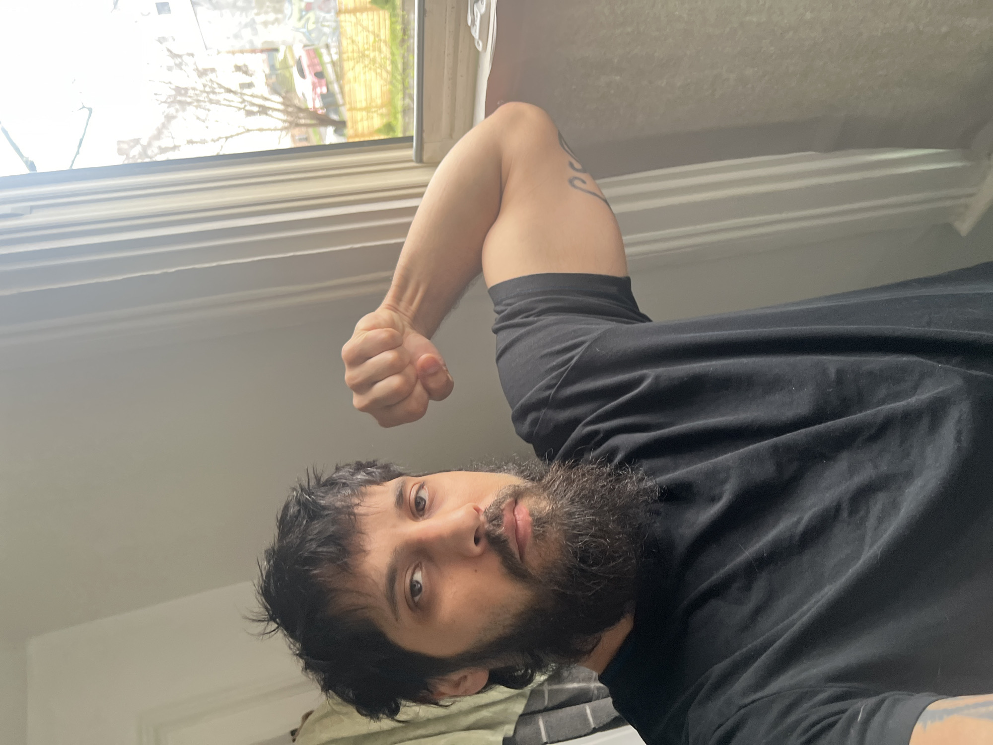 A medium shot, slightly rotated, depicts a person with a dark beard, wearing a black t-shirt, with their arm raised in a clenched fist. A tattoo is visible on their upper arm. The background features a window with white trim and a view of trees and buildings, suggesting an urban environment. Considering that the location provided is City of Newburgh, United States of America, the depicted environment may be present there.