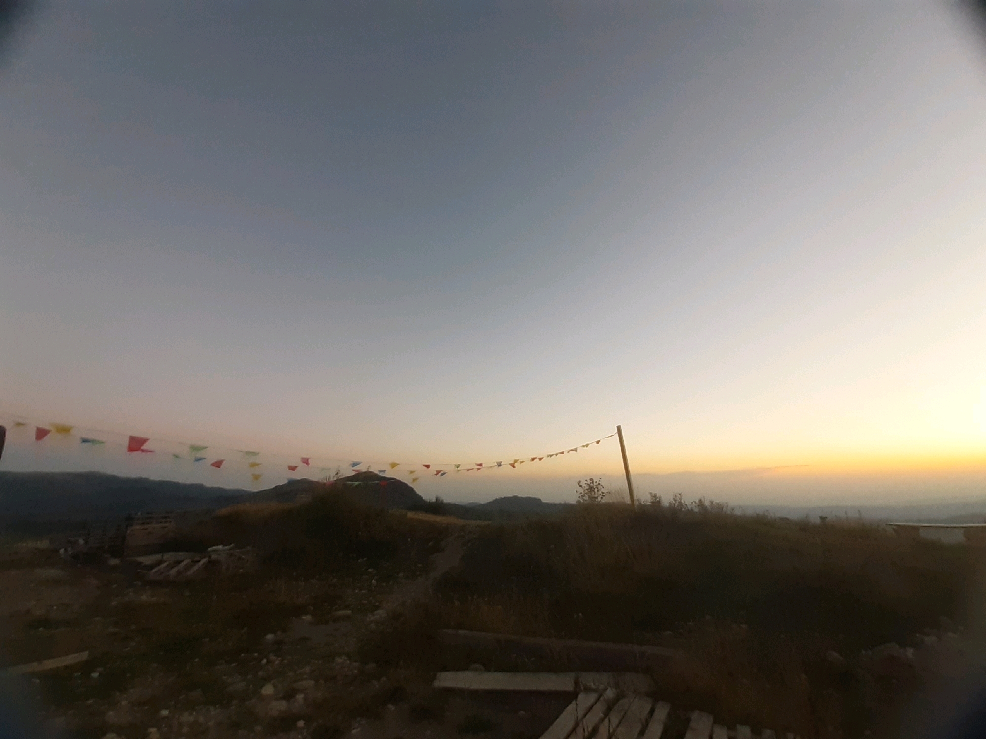This is a wide-angle, horizontal shot of a landscape with a gradient sky, taken at dusk. The sky transitions from a muted grey at the top to a pale yellow near the horizon. A string of colorful triangular flags, primarily red, yellow, and blue, is strung across the middle ground, attached to a wooden post on the right. The flags appear to be a decoration for an outdoor event or gathering.
The foreground is a rocky, uneven terrain with dry grass and some wooden structures. In the midground, there is a series of rolling hills and a darker, more densely vegetated area that obscures the base of the hills. In the far background, mountain ranges are visible, silhouetted against the twilight sky. The overall impression is a rural or semi-rural setting, possibly a park or a viewpoint area. The fisheye lens effect distorts the edges of the image, curving the horizon slightly. The location is identified as Verona, Georgia. There are no people or animals visible in the image.