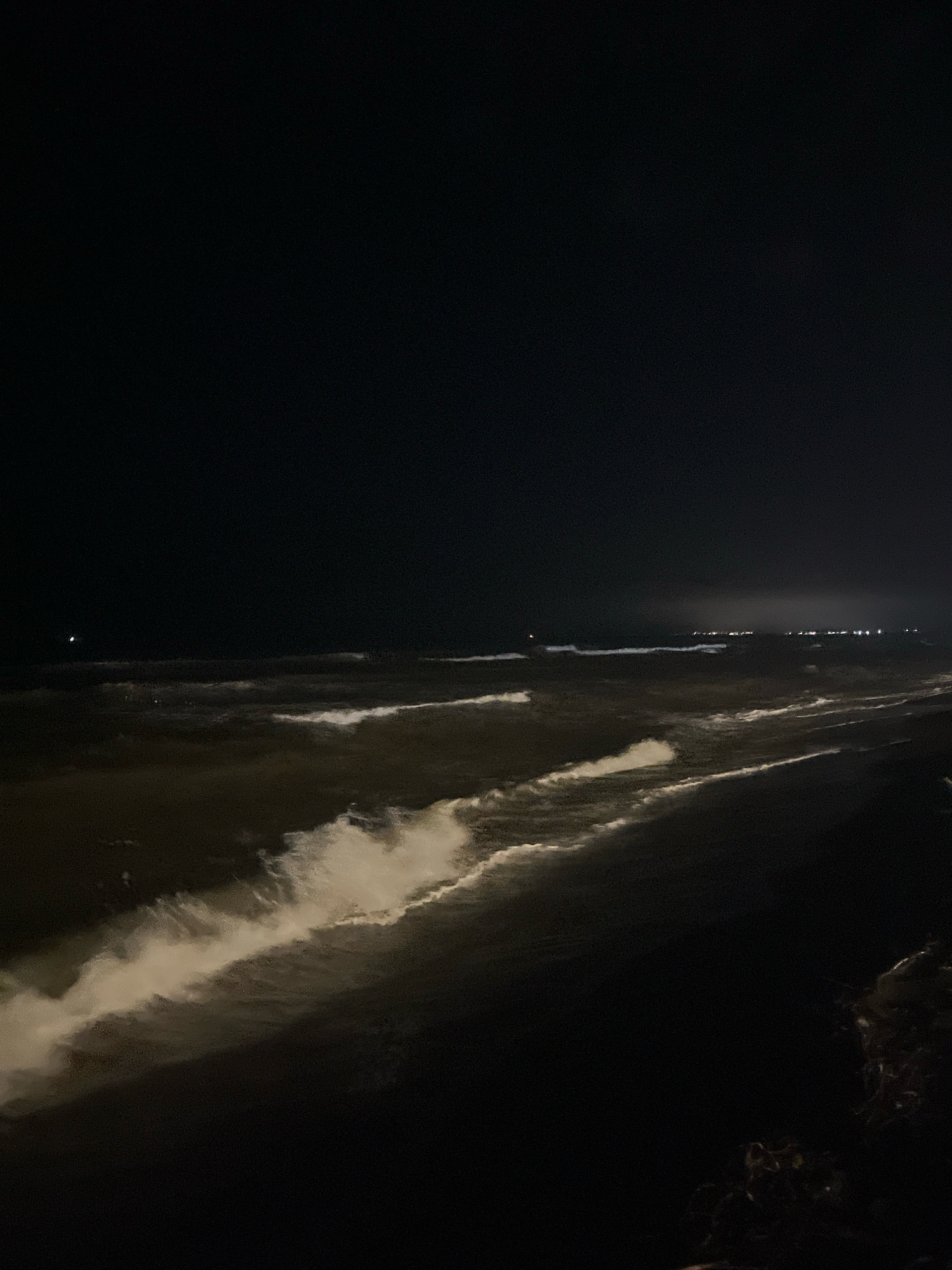This is a night-time, low-angle shot of the Black Sea coast in Ureki, Georgia. The image is predominantly dark, with the exception of the foamy white crests of waves crashing onto a dark, wet sand beach. In the distance, faint lights are visible on the horizon, suggesting a populated area or possibly ships. The sky is a deep black, with no stars or moon clearly discernible. The overall impression is one of a calm but powerful natural scene at night.