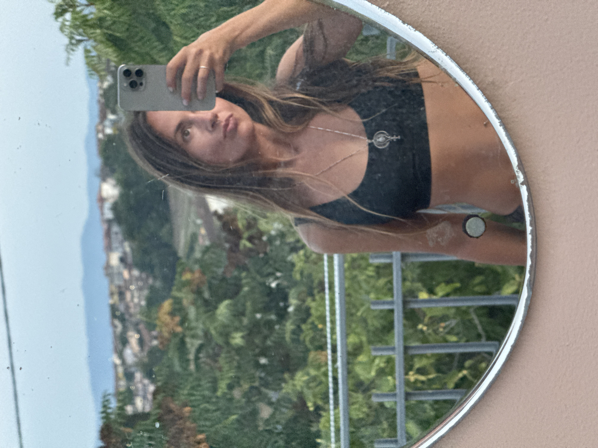A woman with long, light brown hair takes a mirror selfie. She is wearing a black sports bra and a silver necklace with a circular pendant. The mirror is oval-shaped and set in a pale pink frame. The reflection shows a balcony railing and lush green trees behind her, with buildings and mountains visible in the distance. The sky is overcast.