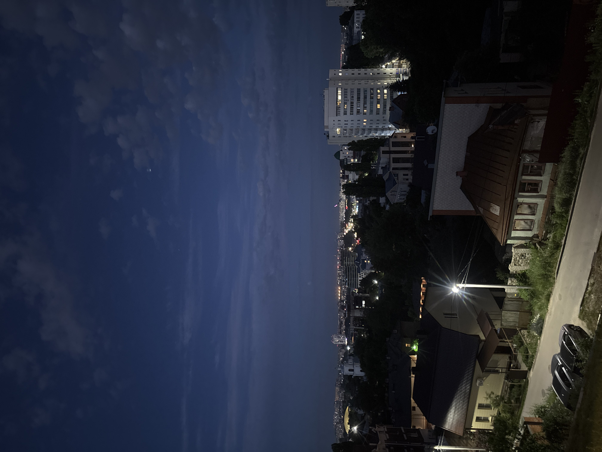 An aerial, low-light, long-shot, vertical photograph captures a nighttime cityscape. In the foreground, a paved street with two parked cars sits to the left. A light pole with an illuminated lamp stands tall on the right side of the street. Beyond the street, a two-story house with a dark, gabled roof and a light-colored exterior is visible. Another building with a red, gabled roof and a white brick facade is partially visible in the mid-ground, slightly to the right.
In the background, a large, multi-story building with many illuminated windows dominates the scene. Further back, the city extends into the distance with numerous lights indicating buildings and streetlights. The sky above is a deep blue with scattered clouds, and a faint crescent moon is visible. The image appears to be taken from an elevated position, possibly a building or hill, looking out over the urban landscape of Novaya Usman', Russia.