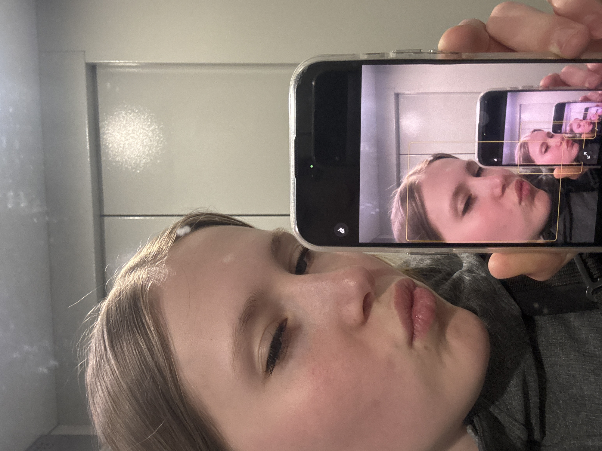 A person, likely female, is taking a selfie with an iPhone. The phone is held in the upper right of the frame, displaying a live view of the same person, recursively. The person has light skin and light brown hair, and is making a kissing face. Part of the person's gray shirt is visible. The background consists of a white wall or door with horizontal lines. There is a round area with white spots on the wall. The image does not contain information to suggest a location in Chaikovsky, Russia.