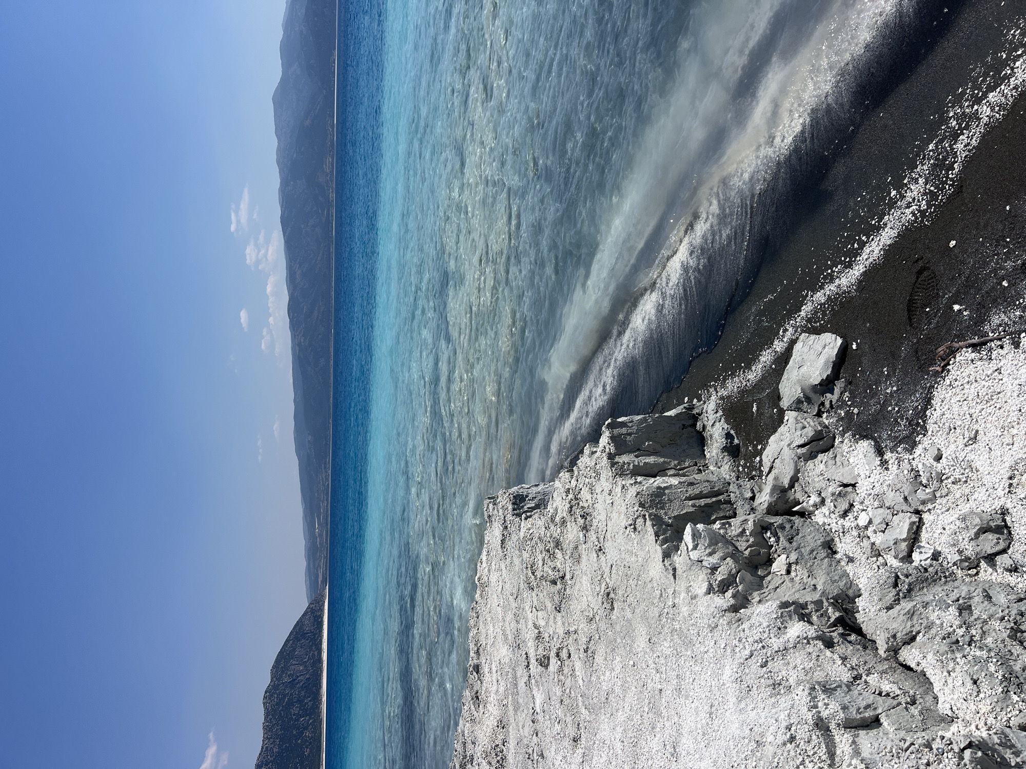 A vertical, eye-level, medium shot captures the edge of a white, rocky cliff overlooking the turquoise waters of Salda Lake in Yeşilova, Turkey. The lake's water transitions from a bright turquoise in the foreground to a deep blue in the background, with a lighter, almost white, sandy shore lining the water's edge. Distant, hazy mountains form the horizon under a clear, pale blue sky with a few wispy white clouds. The rocky cliff face in the foreground is composed of light-colored, layered sediment, with some darker sand and small white pebbles scattered at its base. A single footprint is visible in the dark sand near the water. The overall scene is bright and sunny, with a sense of natural beauty and tranquility.