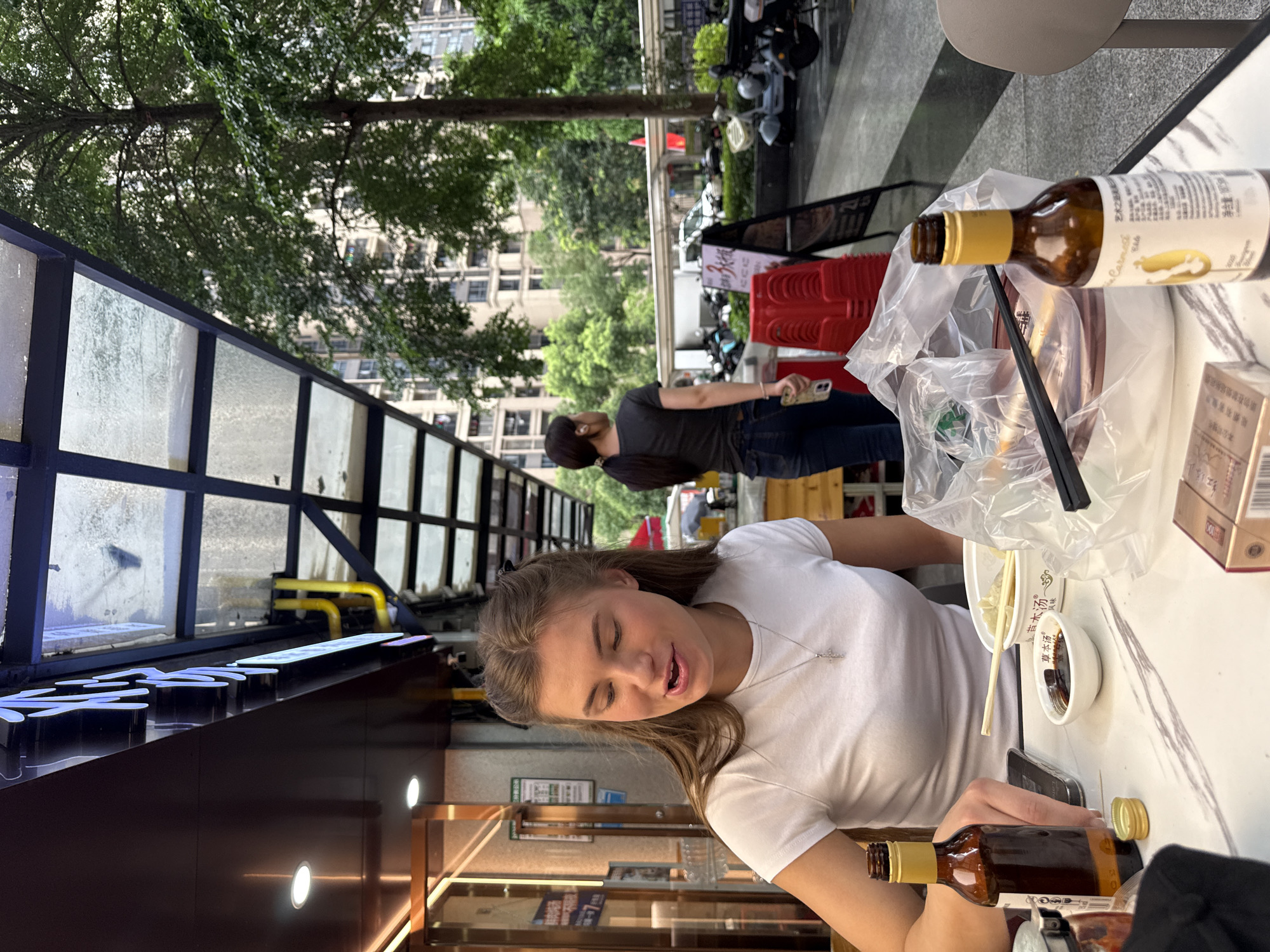 A medium shot captures a young Caucasian woman with long, light brown hair, wearing a white t-shirt and a silver pendant necklace, sitting at a table outdoors. She is looking to her left and speaking. On the table, a brown glass bottle with a yellow label, a small white bowl with dark liquid, a plastic bag containing food and chopsticks, and a pack of cigarettes are visible. A second identical bottle is to her right. Behind her, a woman with dark hair pulled back in a ponytail, wearing a dark t-shirt and jeans, is walking away from the camera. The scene appears to be in Haizhu District, China, indicated by the signage in Chinese characters and the architectural style of the buildings in the background. The environment is a street-side dining area with a covered walkway overhead and lush green trees and modern buildings visible in the distance.