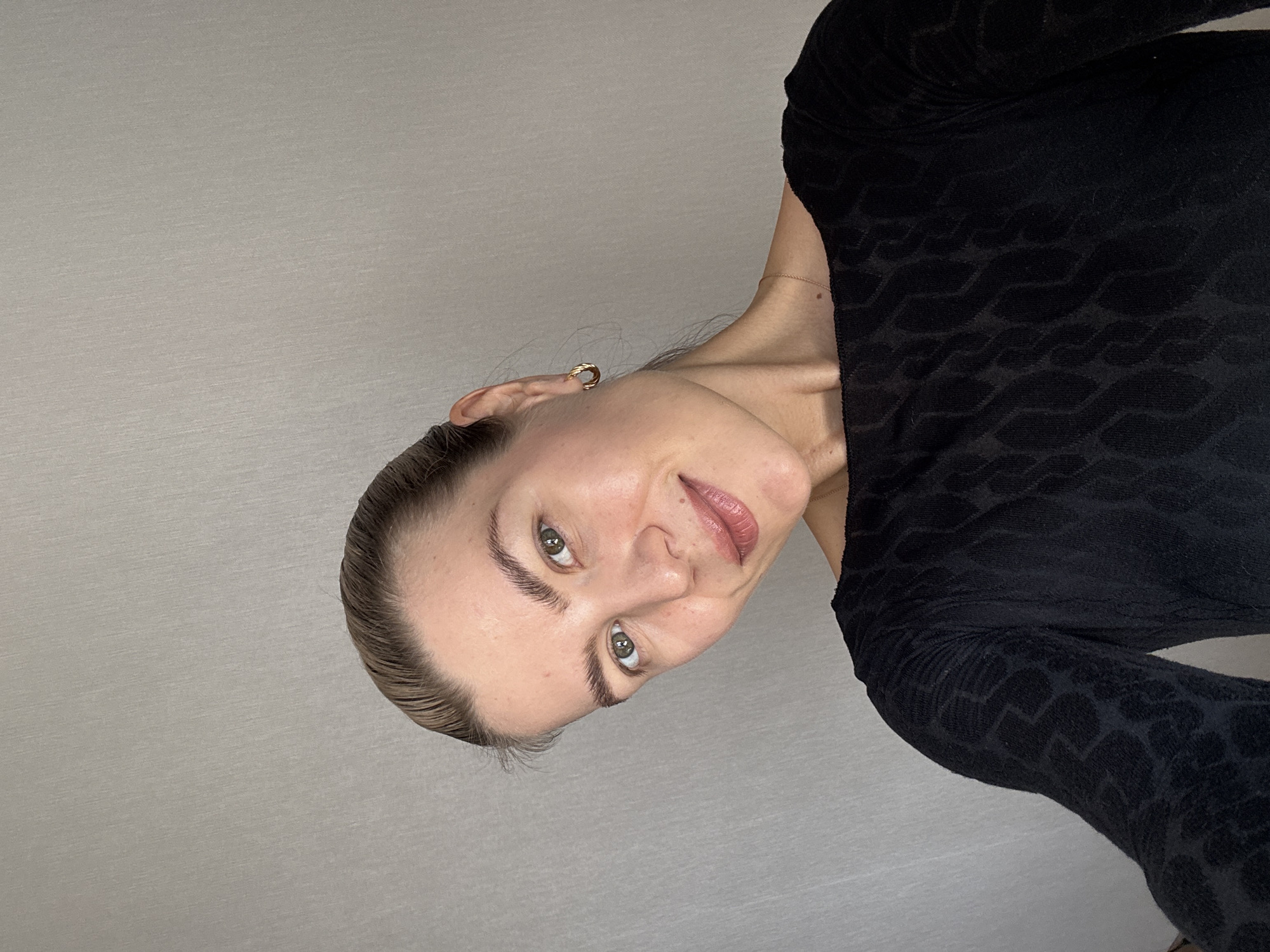 A close-up, rotated portrait shows a woman against a plain, light gray wall. The woman, appearing to be in her 20s or 30s, is positioned as if lying down or tilted significantly, looking towards the viewer with her head tilted back. She has fair skin, light brown hair pulled back from her face, and striking green or hazel eyes with well-groomed eyebrows. A subtle smile plays on her lips, which are colored with a light pink lipstick. She wears a small, twisted gold hoop earring in her visible ear and a very thin gold chain necklace. Her attire consists of a long-sleeved black top that features a subtle, dark-on-dark textured or embossed pattern resembling waves or abstract, organic shapes. The lighting is bright and even, suggesting an indoor setting. There is no visible text.