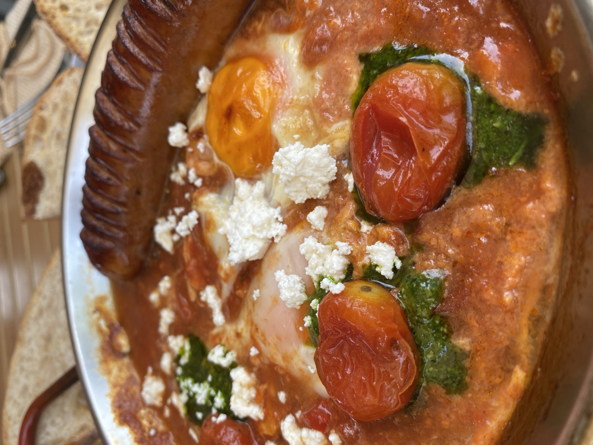A close-up shot of a metal pan filled with a meal consisting of sausage, eggs cooked in a tomato-based sauce, cherry tomatoes, pesto, and crumbled feta cheese. A piece of bread is located on the edge of the plate. The location is Choeng Thale, Thailand.