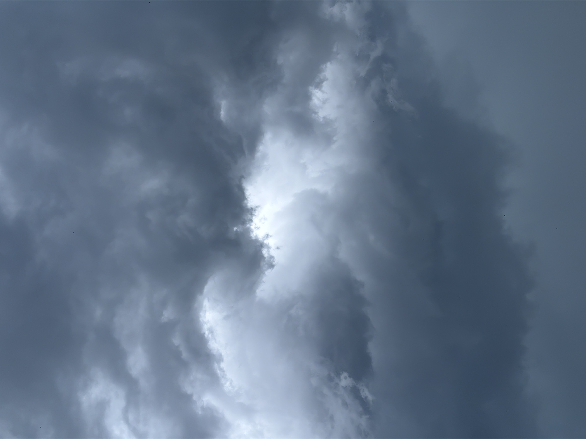 This media file captures a dramatic sky filled with dark, turbulent storm clouds. The clouds are dense and heavy, casting a somber, moody atmosphere. A bright, luminous patch of light breaks through the center, creating a stark contrast with the surrounding darkness. This light illuminates the edges of the clouds, highlighting their textured, swirling forms. The overall impression is one of impending weather, with the light suggesting a break in the storm or the setting sun trying to pierce through the overcast sky. The image does not contain any people, objects, or text. The location and time of day are unknown, but the weather is clearly stormy. The scene evokes a sense of awe and perhaps a touch of foreboding.