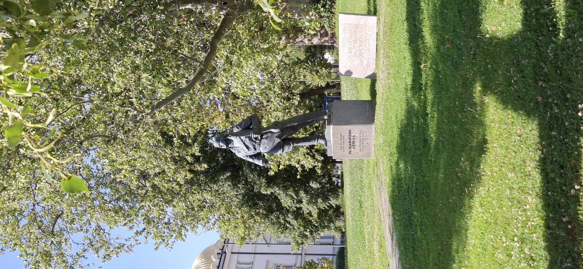 A bronze statue of a male figure in pilot attire stands on a stone pedestal in a grassy park. The statue is the primary subject, depicted from the waist up, with the figure holding a propeller blade. Behind the statue, a large, leafy tree dominates the background, with a clear blue sky visible through its branches. To the left of the statue, a white building with a golden dome can be seen. To the right of the statue, on the grass, is a flat stone monument with inscriptions. The ground is covered in green grass with scattered white clover flowers. The scene is illuminated by natural sunlight, casting shadows from the trees. This is a monument to Bulgarian pilots.