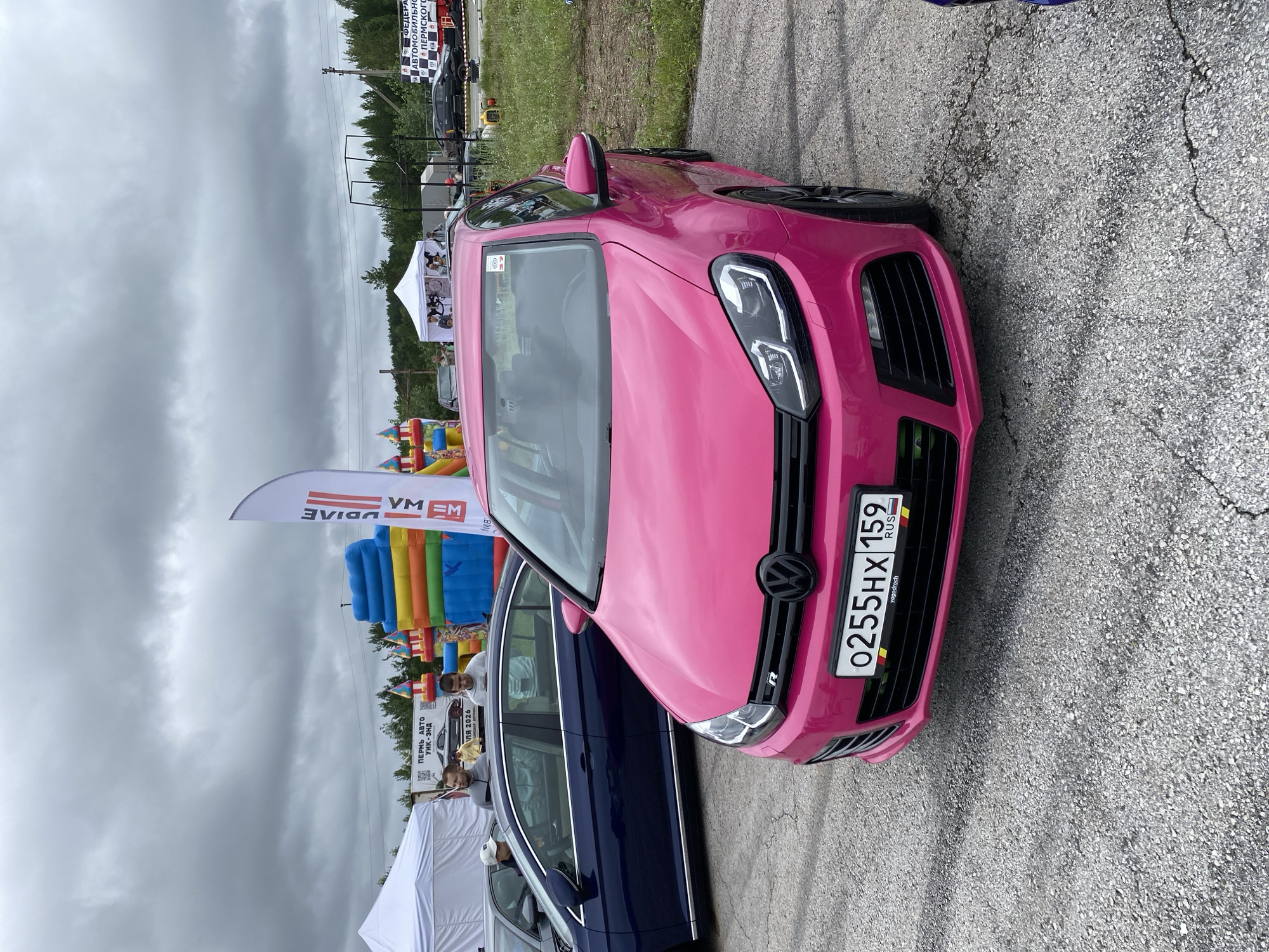 A bright pink Volkswagen Golf R is parked on an asphalt surface, likely a parking lot or road, with a predominantly gray, overcast sky above. The car's front license plate reads "O255HX 159 RUS" and features the Russian flag colors on the right side. In the background, a dark blue sedan is visible, and further back, there are white tents, inflatable play structures (including a colorful castle and slides), trees, and a distant banner that appears to be for "FEDERATSIYA AVTOMOBILNOGO SPORTA PERMSKOGO KRAI" which translates to "Automotive Sports Federation of Perm Krai." This suggests the location is likely an event venue or racing circuit in the Perm Krai region of Russia, possibly related to motorsport or a public event. There are several people visible, some standing near the blue car and others milling around the tents and inflatable structures. The overall scene conveys an outdoor event atmosphere.
