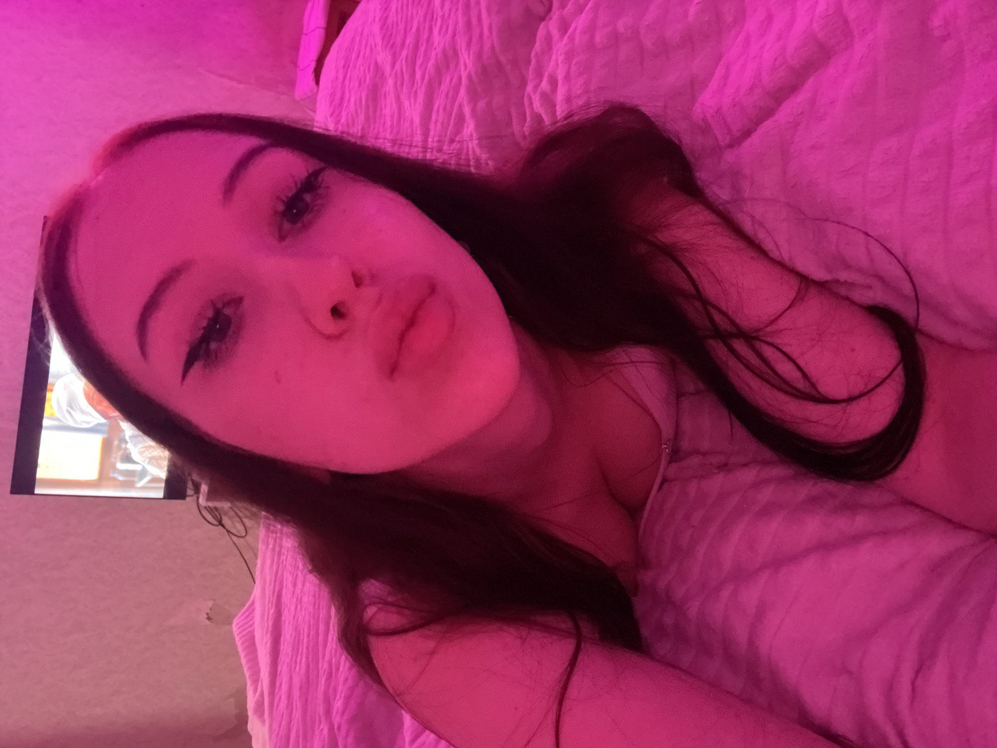 The image shows a young woman lying on a pink bed in a dimly lit room. A television is visible in the background. The lighting in the room is predominantly pink, casting a warm glow over the scene. The woman is looking towards the camera with a pout. Her hair is long and dark, framing her face. She is wearing a light-colored top. The overall impression is one of a casual, personal moment captured in a bedroom setting. There is no discernible information within the image that allows for the identification of a specific location in Votkinsk, Russia.
