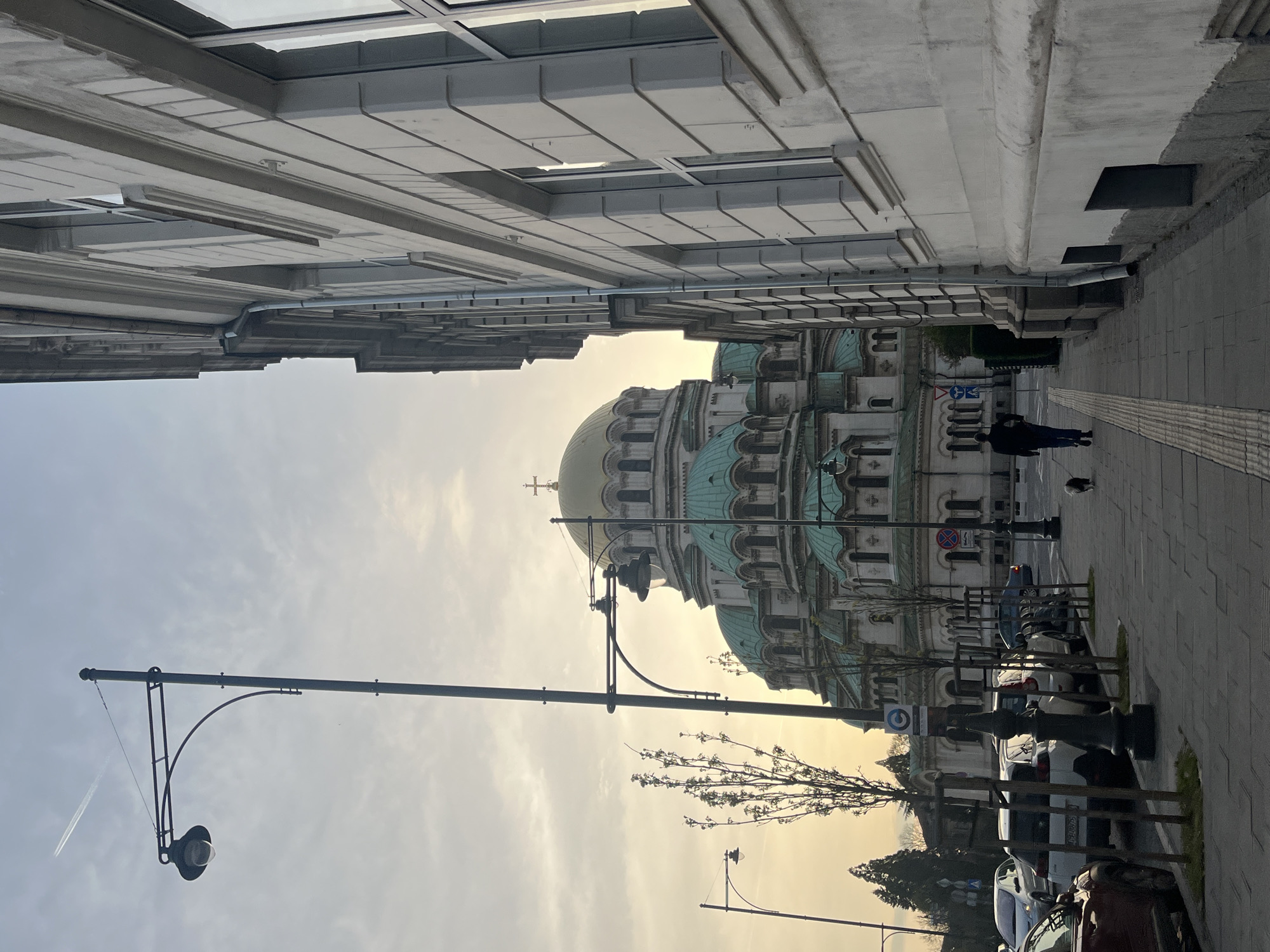 The image shows a street scene in Sofia, Bulgaria, featuring the Alexander Nevsky Cathedral. The cathedral's domes and architectural details are visible in the background. A pedestrian and a small dog are walking on the sidewalk. The street is lined with parked cars and young trees. Streetlights are present. Buildings frame the view of the cathedral, suggesting the perspective of someone standing on the street. The sky is overcast, giving the scene a muted, golden light.