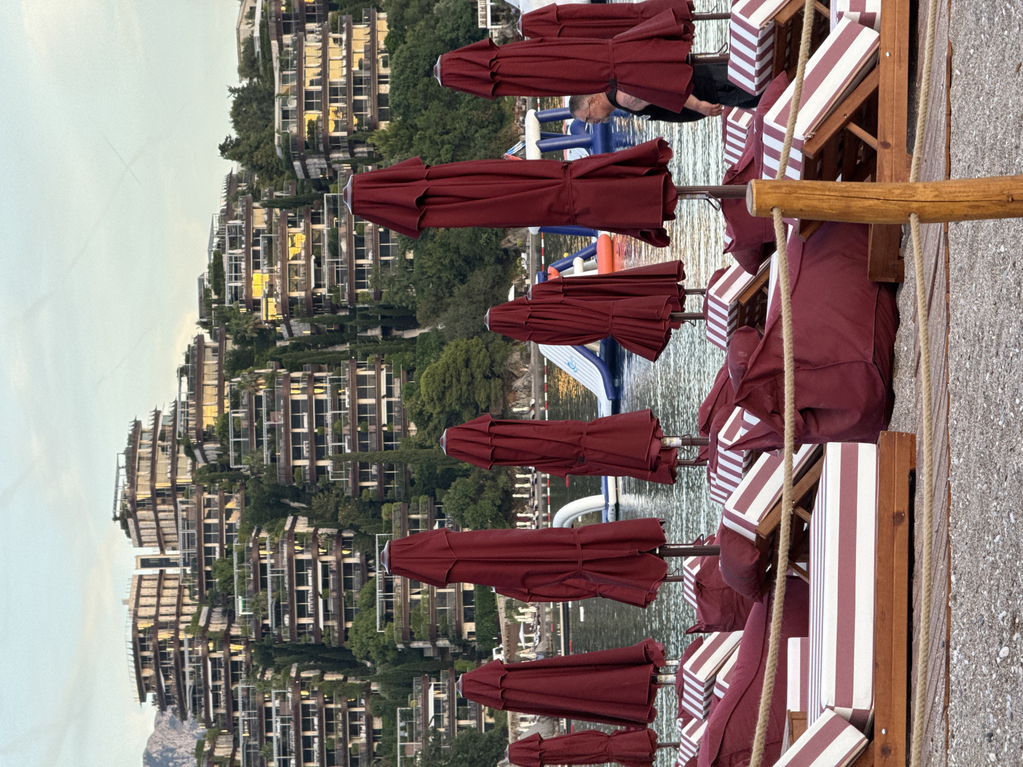 The image depicts a beach scene in Budva, Montenegro, with multiple rows of closed red umbrellas and lounge chairs in the foreground. The lounge chairs feature striped cushions in red and white. In the middle ground, the calm water of the sea is visible, with a few inflatable water toys further out. In the background, a large, multi-tiered resort complex is built into a hillside, characterized by its modern architecture, extensive balconies, and lush greenery. A man is standing to the right of the frame, facing away from the camera, near the water. The sky above is a pale, overcast blue, suggesting it might be late afternoon or early morning.