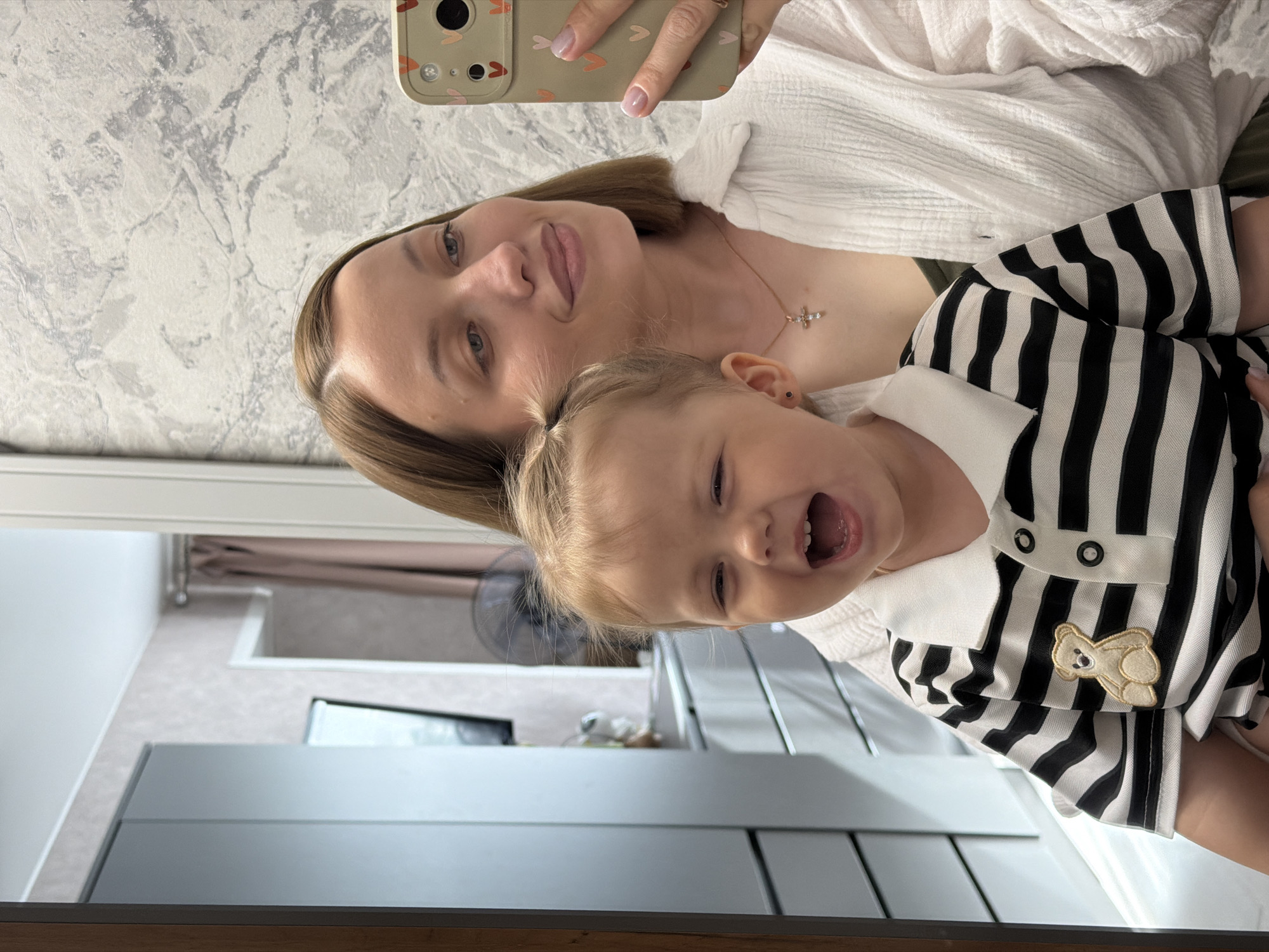 A selfie taken in Reshetnikovo, Russia shows a fair-skinned adult woman with shoulder-length brown hair and a fair-skinned toddler with blonde hair. The woman is wearing a white, textured shirt and a gold necklace with a cross pendant. She is holding a smartphone in her hand. The toddler is wearing a black and white striped shirt with a white collar and a teddy bear patch. The toddler is smiling and open-mouthed. The reflection in the mirror reveals a bathroom with a white vanity and a gray countertop. A shower curtain is visible in the background. The wall behind the woman is gray with a subtle marble pattern.