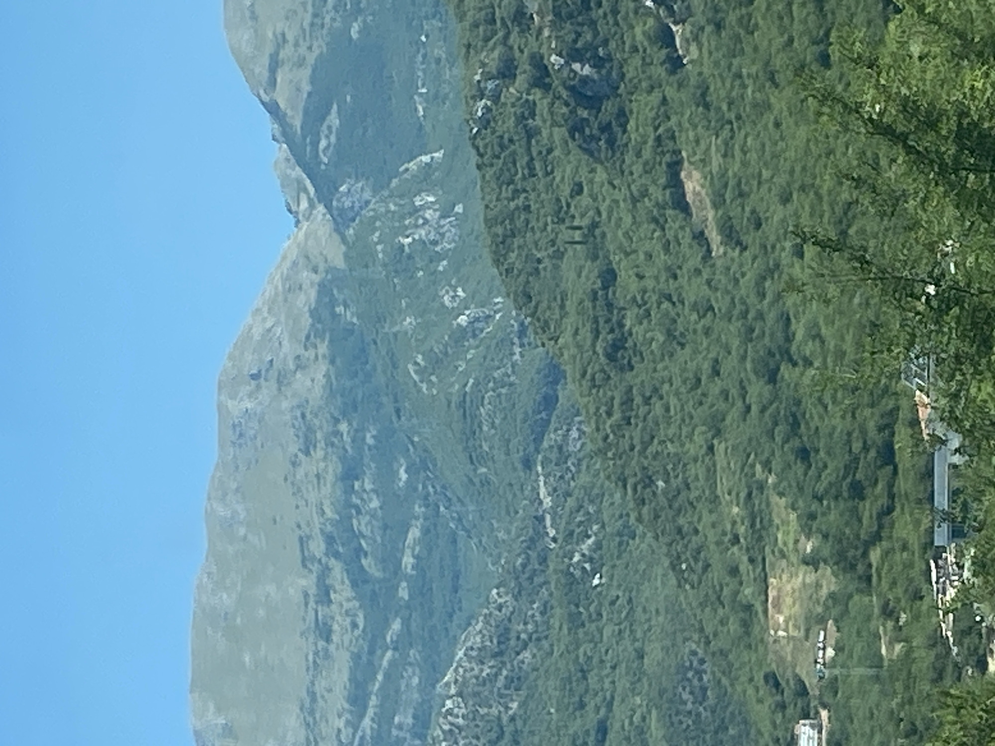 The image depicts a mountainous landscape with a clear blue sky. In the foreground, there are lush green trees, obscuring a part of the view. Further back, rolling hills covered in dense vegetation are visible, with some patches of lighter colored earth or rock formations interspersed. The middle ground shows more rugged mountains with rocky outcrops and less dense vegetation. In the distance, the peaks of larger mountains are visible, their slopes appearing barren or sparsely covered. On the lower left side of the image, a few buildings, likely houses or small structures, are present amidst the greenery, suggesting a rural or semi-rural settlement. The overall scene is characteristic of a Mediterranean mountainous region, possibly the area around Prijevor, Montenegro, known for its scenic natural beauty. There are no people or discernible actions depicted in the image.