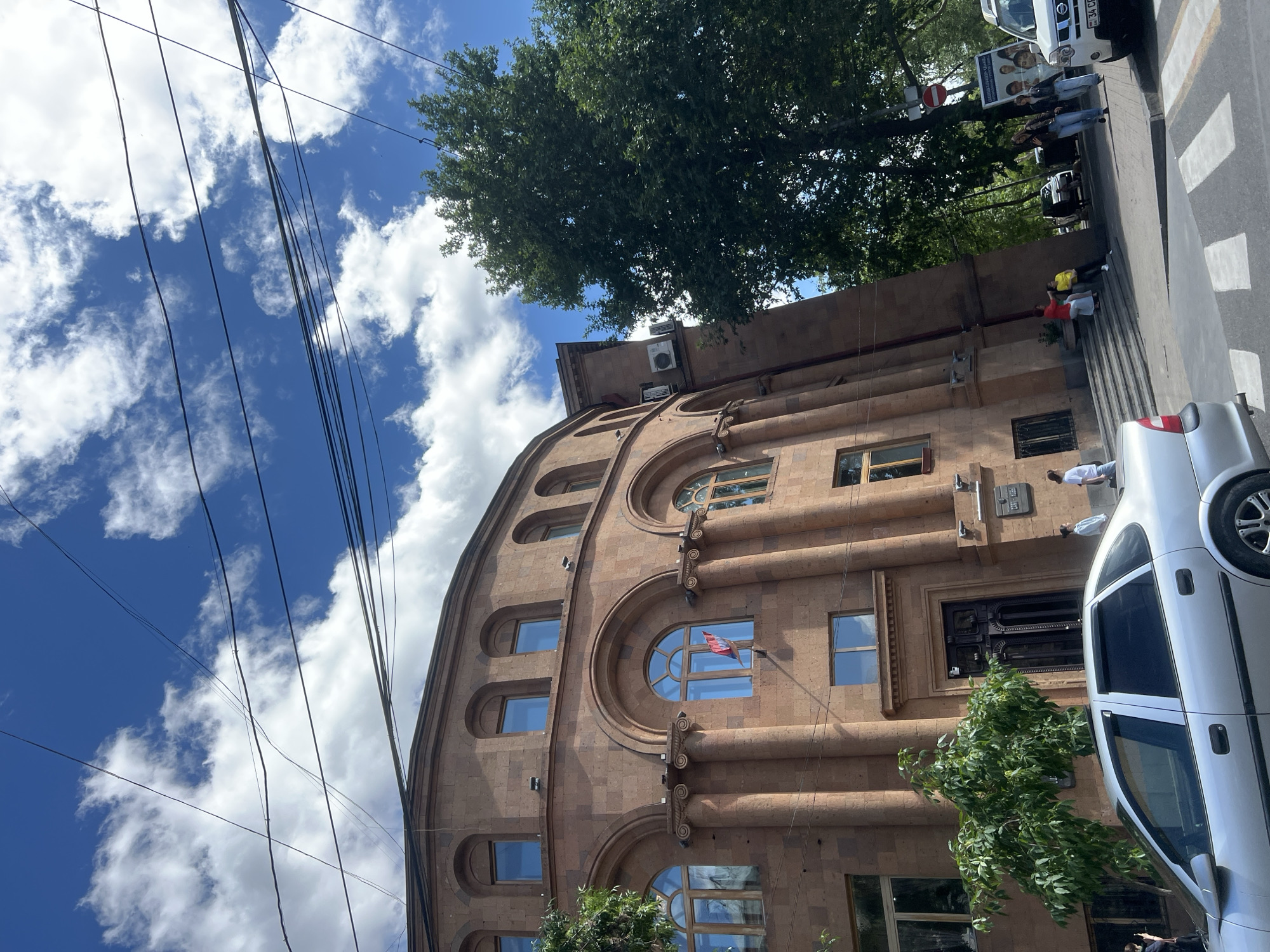 The image shows a portion of a building in Yerevan, Armenia. The building is constructed of reddish-brown stone, with an arched facade and multiple windows. An Armenian flag is visible in one of the arched windows. Power lines cross the sky above the building, which is partially obscured by a tree. A silver car is parked along the sidewalk in front of the building. Pedestrians are visible near the crosswalk and the building's entrance.