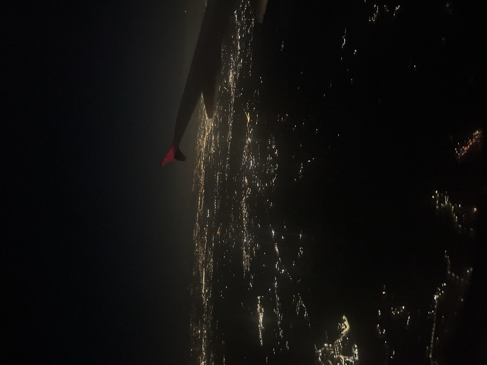 The image depicts an aerial view, likely taken at night, of what appears to be a coastline with scattered lights suggesting a city or town. The lights are a mix of white and yellow, indicating various sources like streetlights and building illumination. In the foreground, a dark shape with a red appendage or marker is visible, possibly part of the aircraft or drone from which the image was captured.
While the details are limited, the general characteristics of the coastline and the scattered lights potentially align with the coastal areas of Trevignano Romano, Italy, as it is situated along the shores of Lake Bracciano. The environment suggests a night-time setting, with an emphasis on the lights illuminating the settlement. Further analysis would be needed to confirm this location.