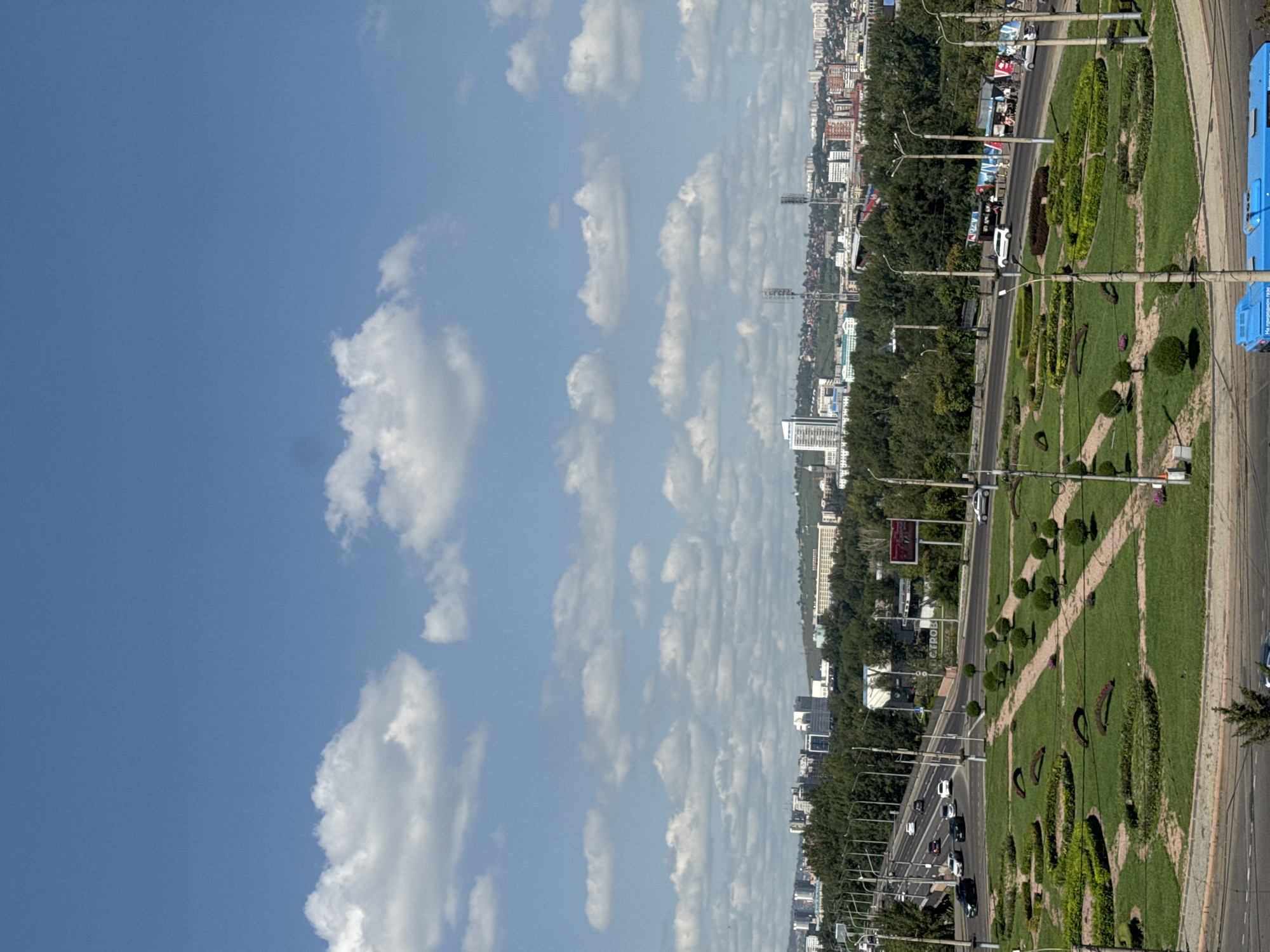 The image captures a panoramic view of a city, likely Krasnoyarsk, Russia, on a bright, partly cloudy day. The sky is a vibrant blue, dotted with fluffy white cumulus clouds. In the foreground, a circular green park area is meticulously landscaped with trimmed bushes, small trees, and walking paths. The park appears to be situated in a roundabout or at an intersection.
Beyond the park, a multi-lane road with moderate traffic is visible. Several cars, including black SUVs and white sedans, are driving on the road, and a blue bus is parked at the lower right edge of the frame. Streetlights and utility poles line the road and park.
In the mid-ground, a dense line of green trees forms a backdrop to the park. Above the trees, a city skyline emerges, characterized by a mix of modern buildings, including high-rise apartments and office buildings. Some of these buildings are white, while others are brick-colored. Further in the distance, rolling hills are visible, suggesting a natural landscape surrounding the urban area. A few tall communication towers are also scattered throughout the city. One billboard with red text is visible on the right side of the road. The overall impression is of a well-maintained urban environment with a blend of green spaces and developed infrastructure.