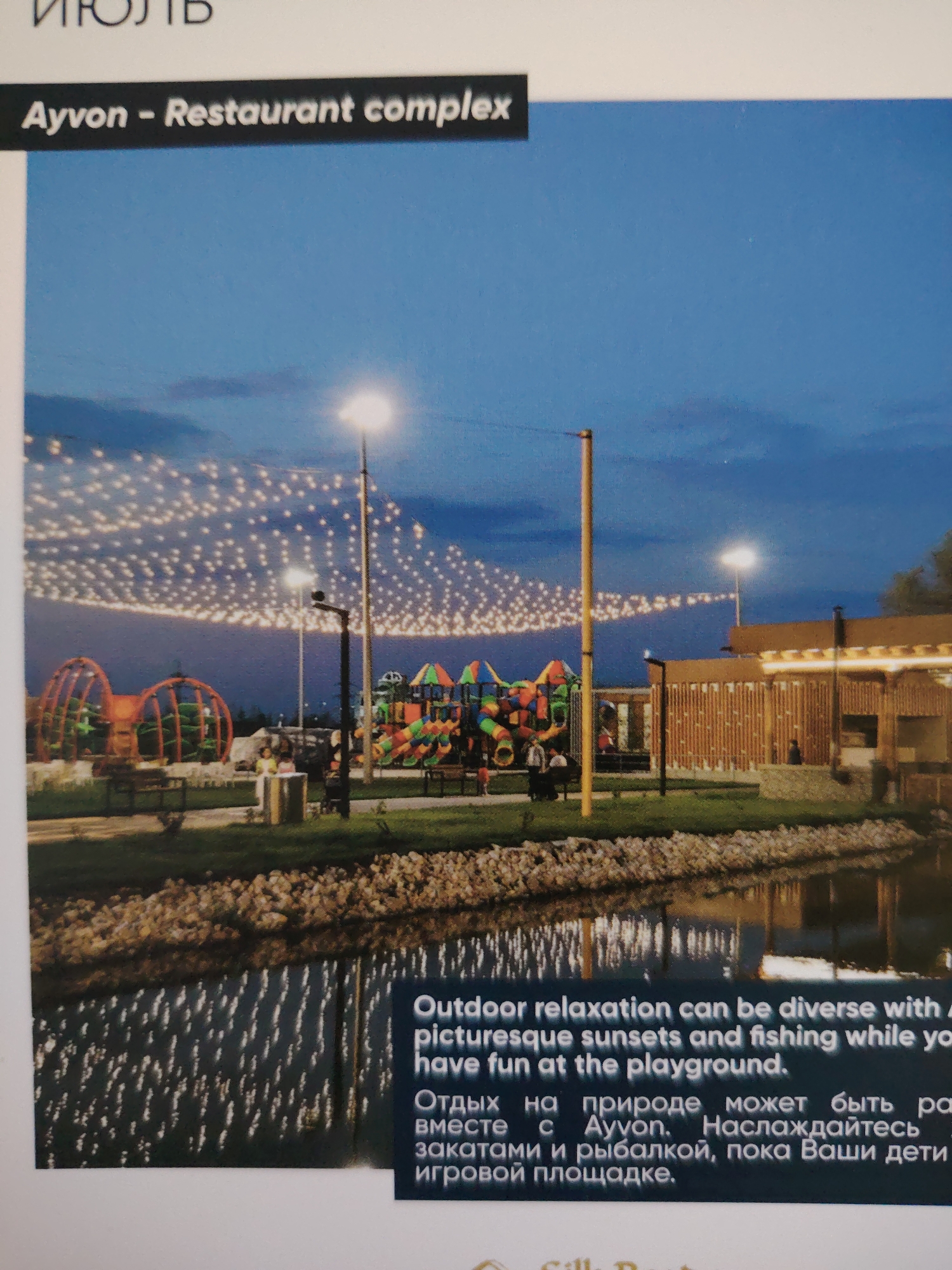 The image depicts an outdoor recreational area at dusk, illuminated by strings of warm-toned fairy lights stretched across the sky and by bright spotlights. In the foreground, a body of water reflects the lights, with a rocky bank separating it from a grassy area. Behind the bank, a playground with colorful slides and structures is visible, along with several people interacting in small groups. To the right, a modern building with a slatted wooden facade and an illuminated awning suggests a restaurant or service area, with a few individuals seated or standing near it. The scene conveys a festive and relaxing atmosphere. The text on the image identifies the location as "Ayvon - Restaurant complex" and includes Russian text that translates to "Outdoor relaxation can be diverse with picturesque sunsets and fishing while you have fun at the playground." There is no information within the image or provided context that places this location in the city of Bahshitpa, Uzbekistan.