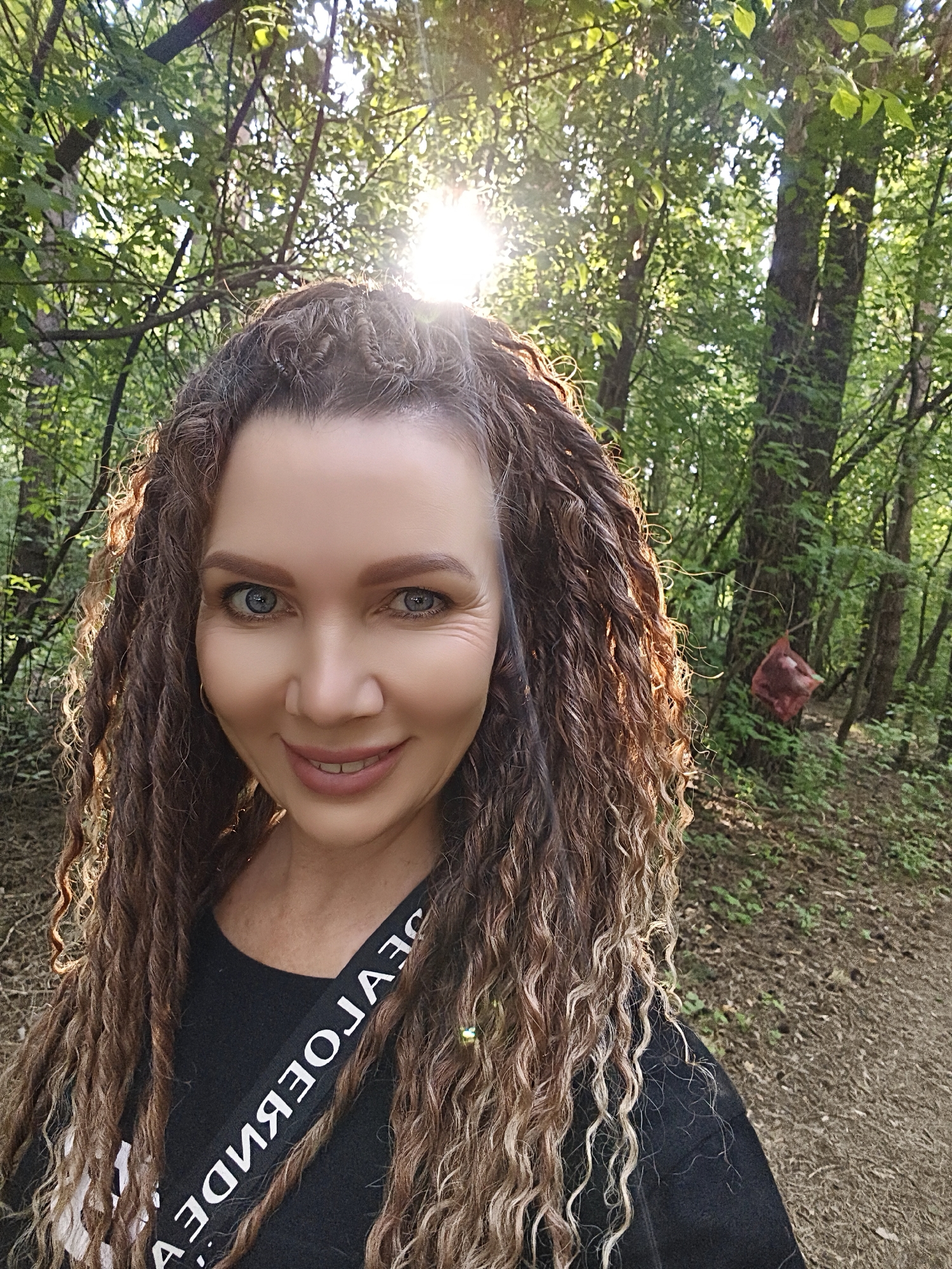 The image depicts a woman with long, curly brown and blonde hair standing on a path in a wooded area. She is wearing a black shirt with a strap across her shoulder. The background consists of trees and foliage, with sunlight filtering through the trees. A red object hangs from a tree in the distance. The location is unspecified.