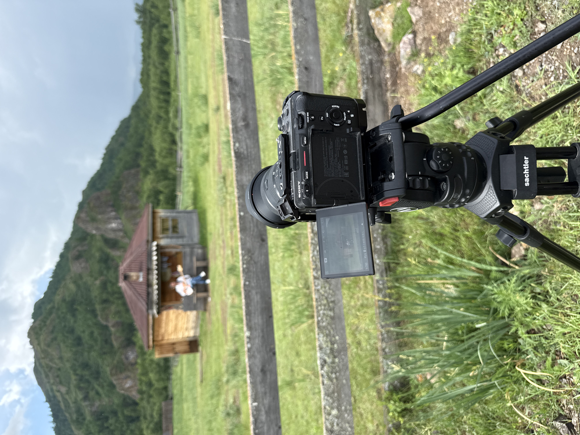 In Anchyl-Chon, Russia, a camera on a Sachtler tripod captures a scene featuring a person playing a guitar in front of a small wooden house. The house has a dark roof and wooden walls, with a porch area where the person is seated on a bench. Behind the house, a lush green mountain range covered in trees rises under a cloudy sky. A weathered wooden fence runs horizontally across the mid-ground of the image, separating the foreground with the camera from the rural landscape. The camera itself is a Sony model with a lens attached, and its screen is visible, displaying the framed shot. The ground is covered in green grass and some wild vegetation.