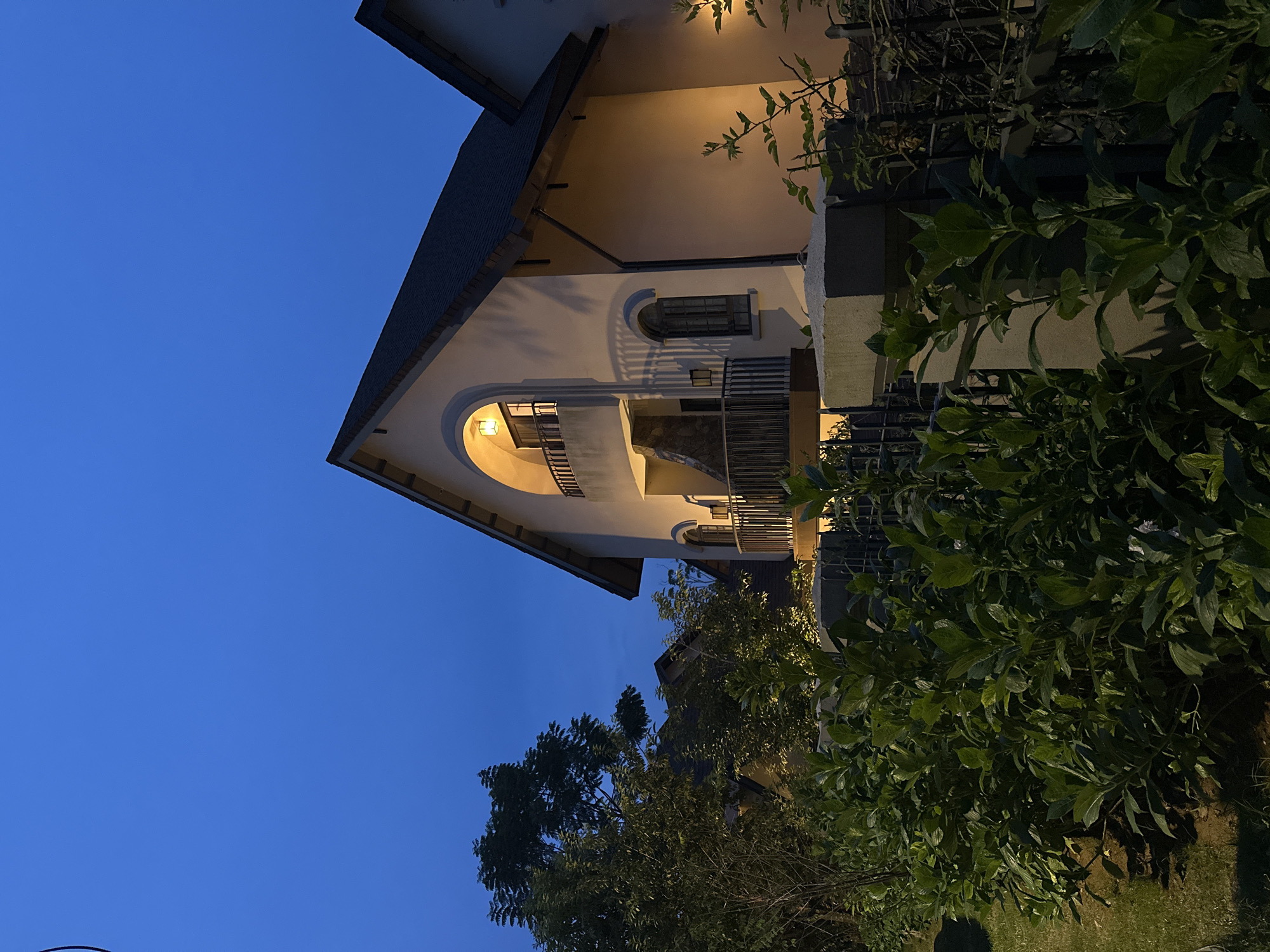 The image captures a nighttime view of a building, likely a house or a small hotel, with a dark blue sky. The building's exterior is illuminated by warm indoor lights, creating a welcoming glow. Architectural details such as arched windows and balconies are visible, suggesting a traditional or classic design. Lush green foliage surrounds the building, with some branches reaching towards the structure. The scene conveys a sense of calm and tranquility, with the soft lighting and natural elements creating an inviting atmosphere. The time of day is clearly night, indicated by the dark sky and artificial lighting. The location context of Lâm Viên - Đà Lạt, Vietnam, hints at a mountainous or elevated setting, possibly with cooler temperatures conducive to evening strolls. There are no people or specific events depicted, focusing entirely on the architectural and natural beauty of the scene.