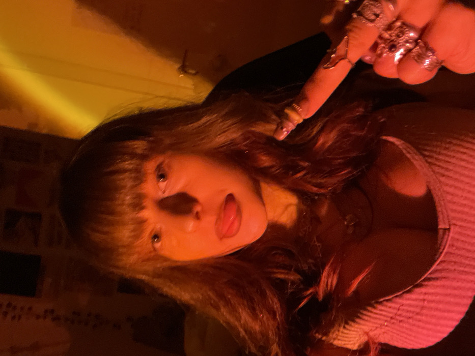 A young woman with bangs sticks out her tongue. She wears several rings on her fingers. Her hair is long and wavy. A gold light washes the scene. She wears a light pink ribbed tank top and a layered necklace. The camera angle is from above.