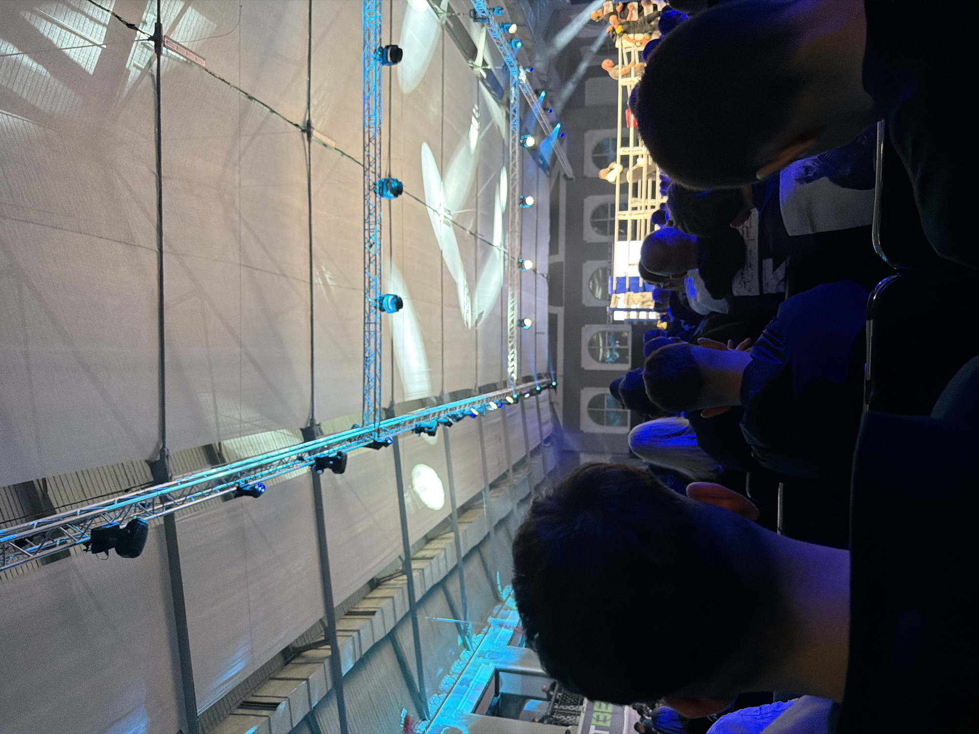 The image shows an indoor event, likely a concert or conference, in Sofia, Bulgaria. The scene is illuminated with bright, artificial lighting, including spotlights and LED bars. Several rows of seated attendees are visible in the foreground. The crowd includes men and women, some with visible faces. The audience is facing towards a large stage area with a backdrop displaying illuminated patterns. Architectural details resembling building facades are partially visible in the background. The overall atmosphere appears to be focused and engaged.