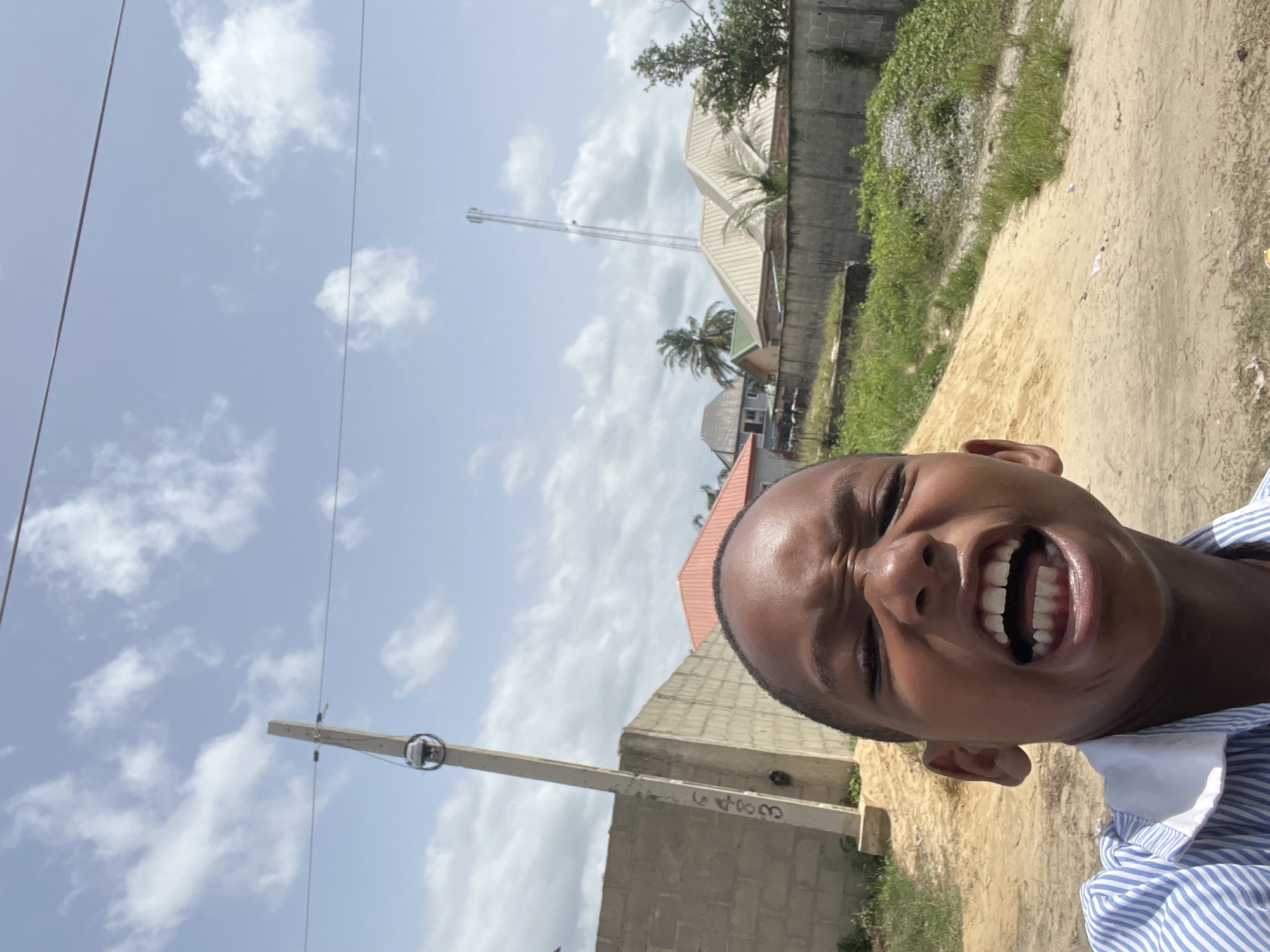 A close-up selfie taken outdoors in Iranla, Nigeria, features a person with dark skin, short black hair, and wearing a blue-and-white striped shirt. The person is smiling broadly, with their teeth visible. In the background, there's a partly cloudy blue sky. Also visible are utility poles with wires, brick buildings, a cell phone tower, palm trees, and vegetation. There is a dirt ground in the foreground.