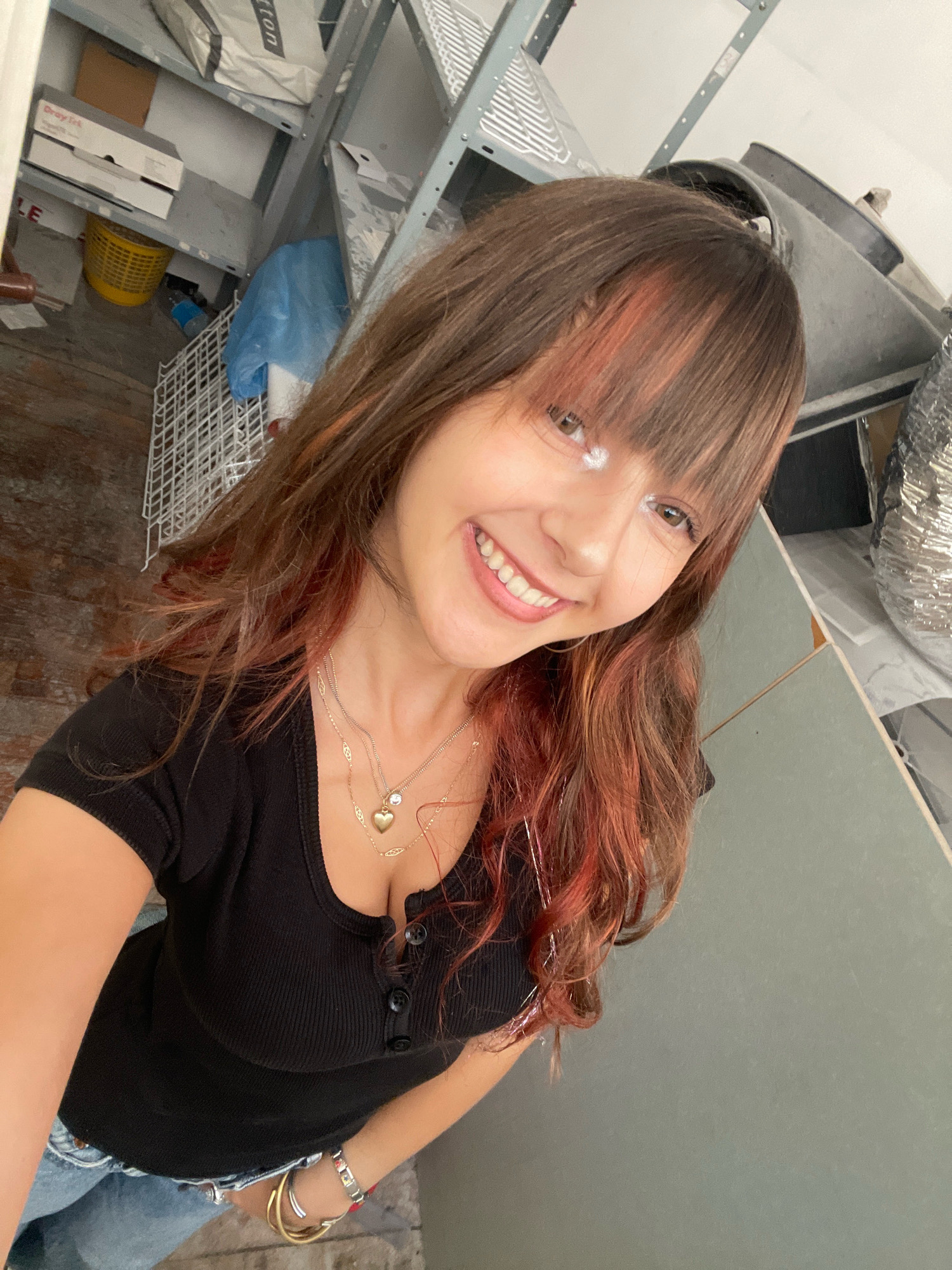 The image shows a young woman taking a selfie in what appears to be a storage area or garage. She is wearing a black, ribbed, short-sleeved shirt with buttons at the neck and blue jeans. She has long, brown hair with reddish highlights and bangs. She is smiling and wearing two necklaces, one with a heart pendant and another with a small clear stone. She also has bracelets on her wrist. The background is cluttered with shelves, boxes, a yellow basket, a blue plastic bag, and various metal objects, including what looks like HVAC ducting. The floor is wooden. There is no specific indication of the location being in Raanana, Israel.